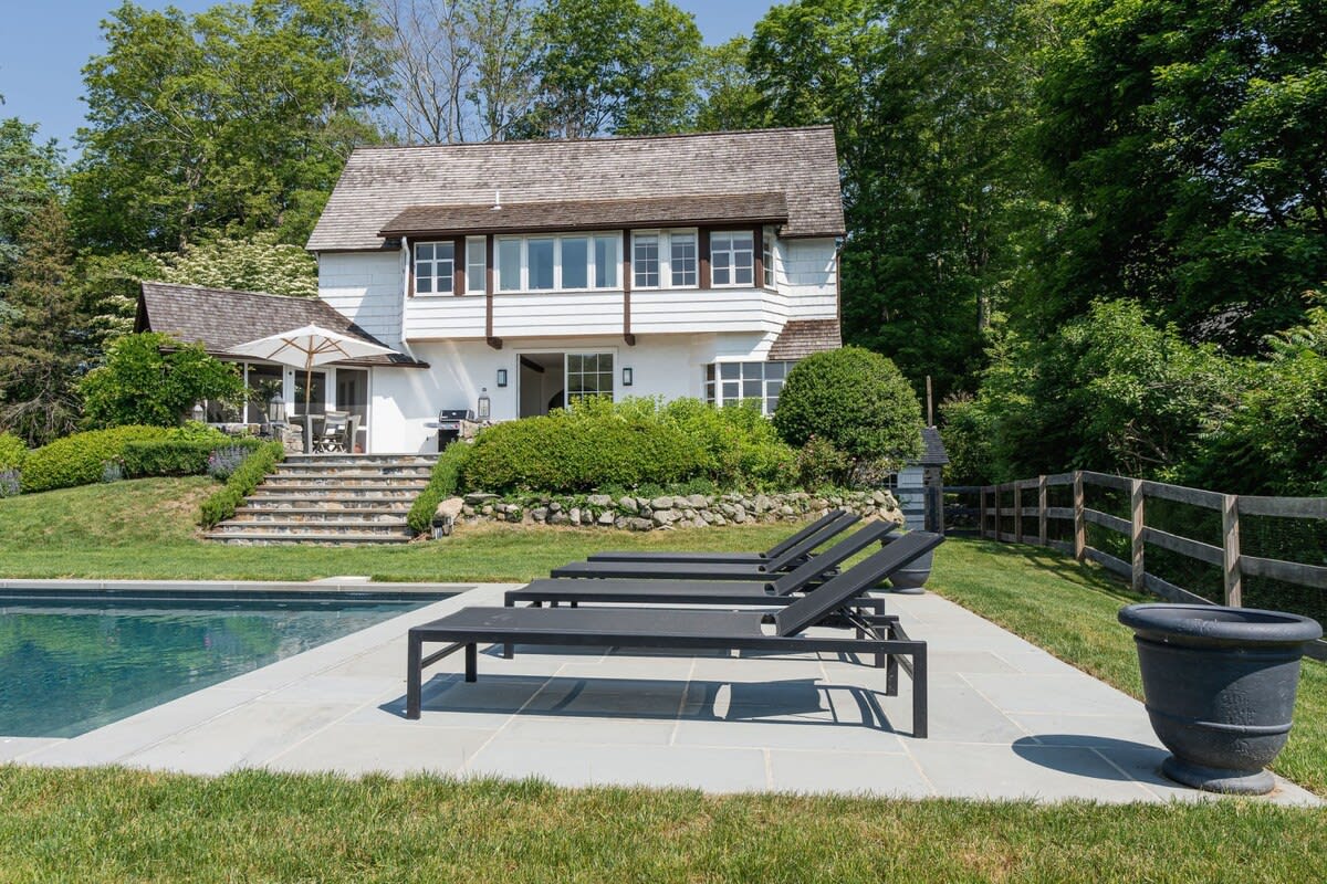 The back of a white two-story house with dark trim and a shingled roof, overlooking a rectangular inground pool. A light grey stone patio next to the pool features four black modern lounge chairs and a large dark planter. Lush green grass and mature trees frame the property.