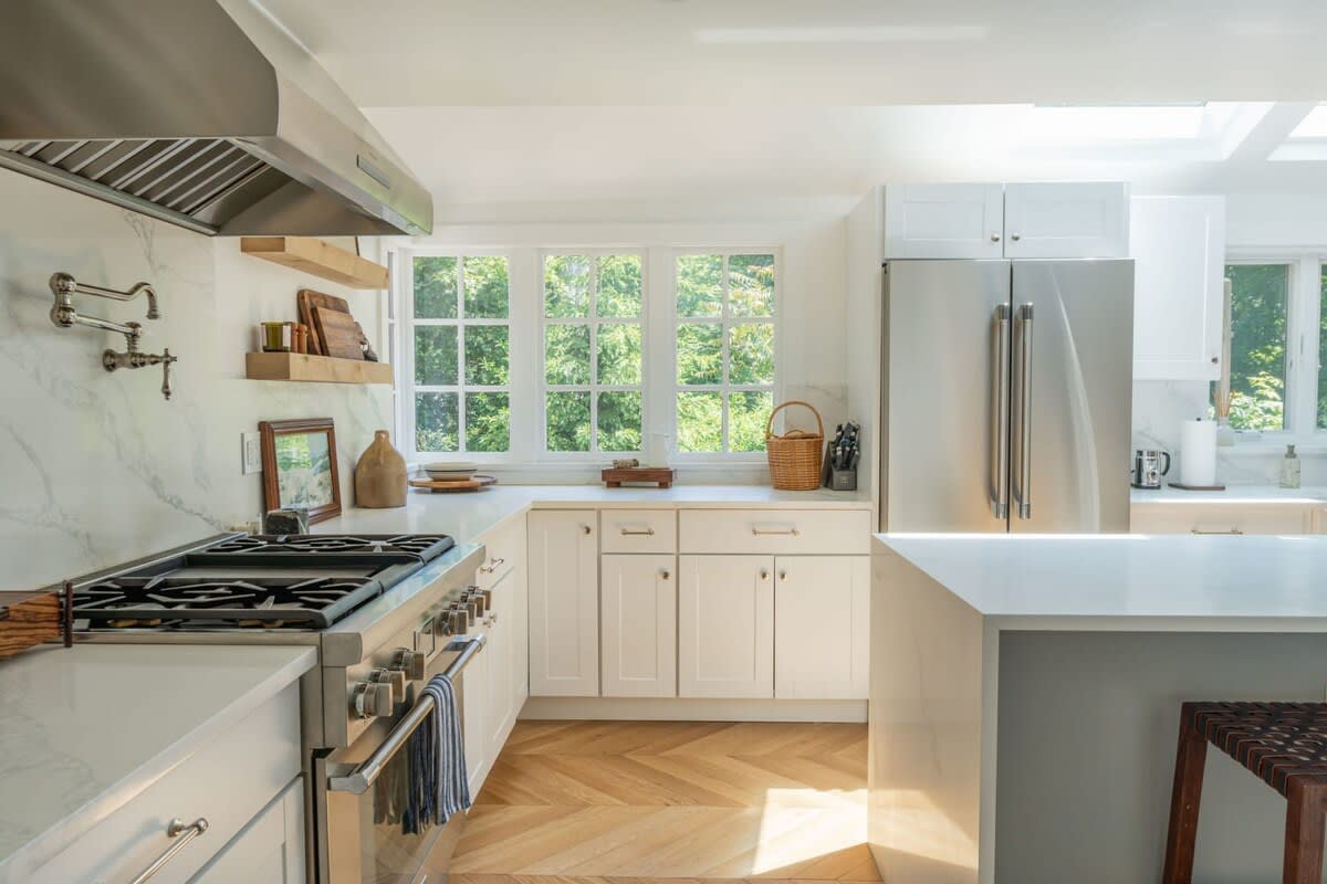 A bright, modern kitchen with white shaker cabinets, light countertops, stainless steel appliances, and a large gas range, featuring a window overlooking green trees and herringbone wood floors.