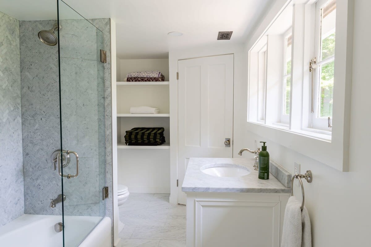 A bright bathroom with white walls and marble-tiled floors features a shower-tub combo with a glass enclosure and herringbone marble tiles. A white vanity with a marble countertop and a large window provides natural light.