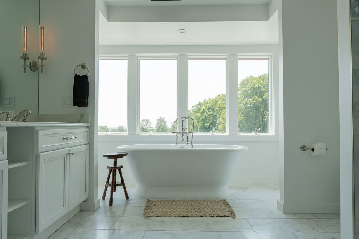 Bright bathroom featuring a white freestanding soaking tub positioned in front of large windows with views of trees. The room has a white tiled floor and a small wooden stool.