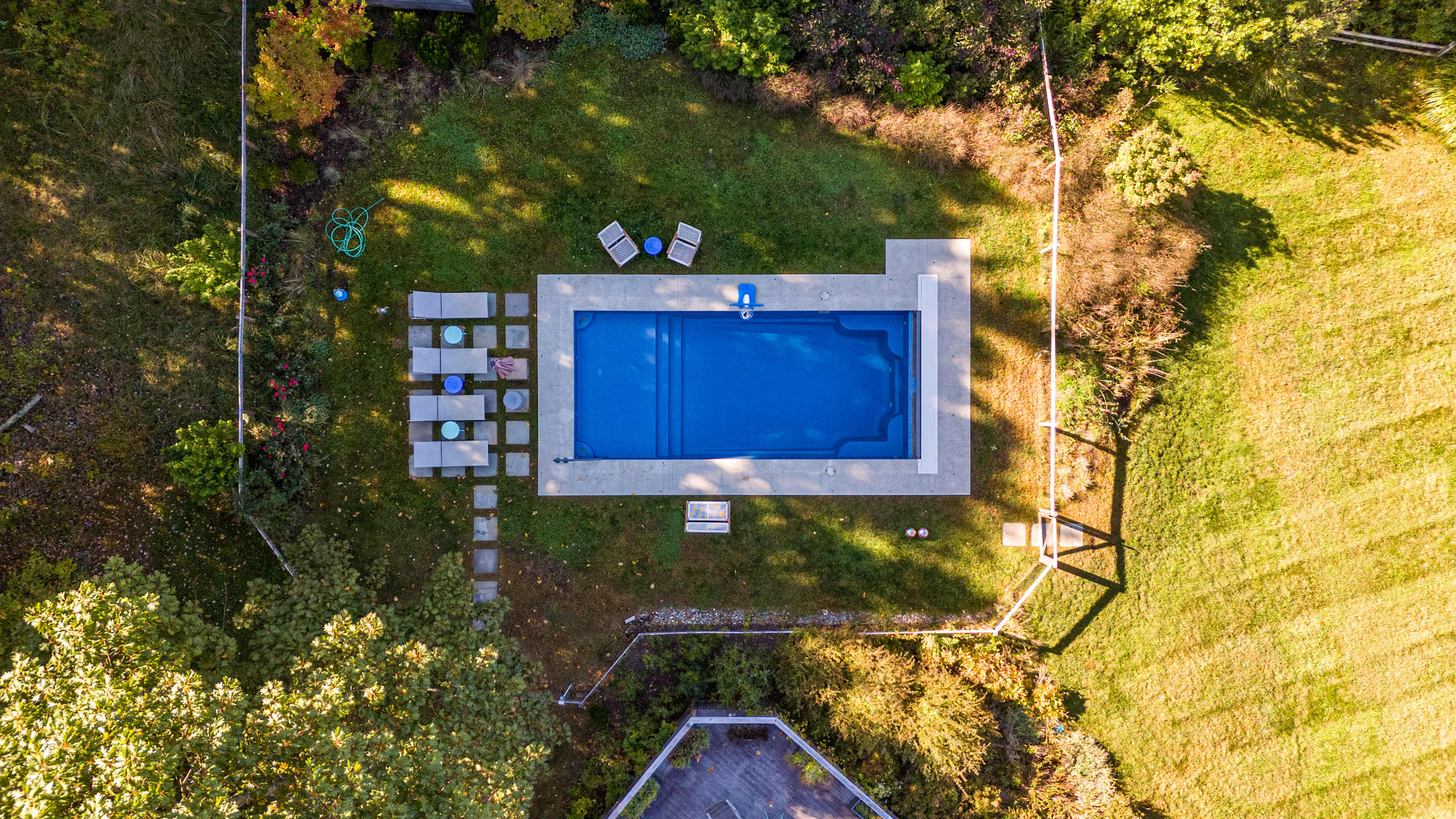 A direct overhead drone view of a rectangular inground swimming pool with a light-colored concrete surround, situated in a well-maintained grassy yard. Patio furniture and stepping stones are arranged around the pool, with the corner of the house visible.