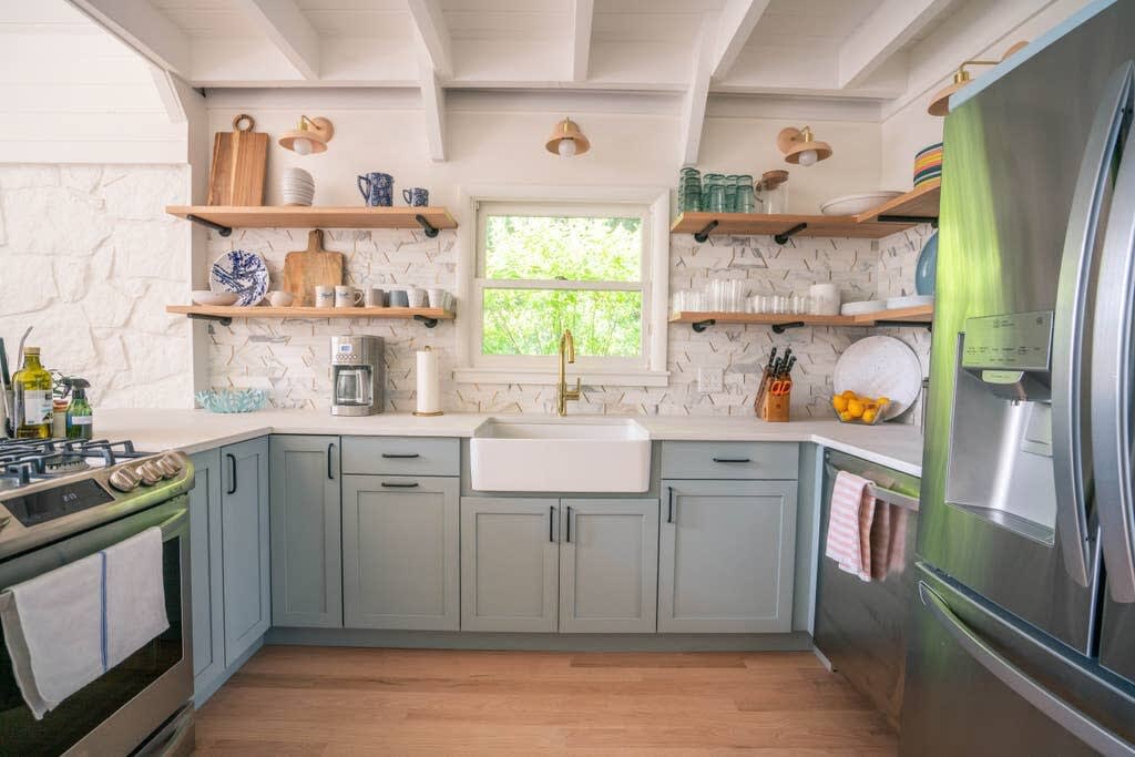 Bright kitchen with light blue shaker-style cabinets, white quartz countertops, a farmhouse sink with a gold faucet, and open wooden shelving against a white geometric tile backsplash. Stainless steel appliances, including a gas range and refrigerator, are visible.