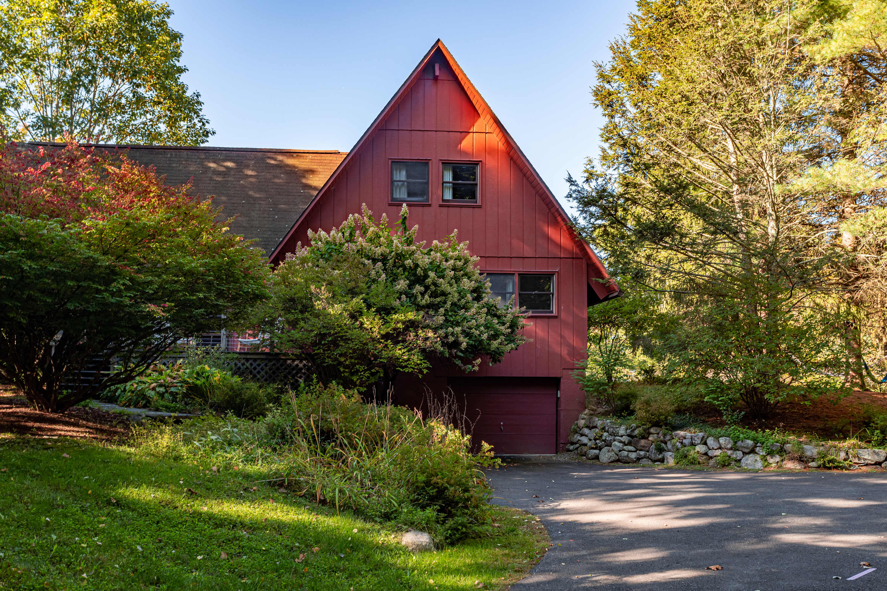 The front facade of a striking red A-frame house with a dark roof, featuring multiple windows and an integrated garage door at ground level. The property is framed by a paved driveway and vibrant green and autumnal shrubs and trees.