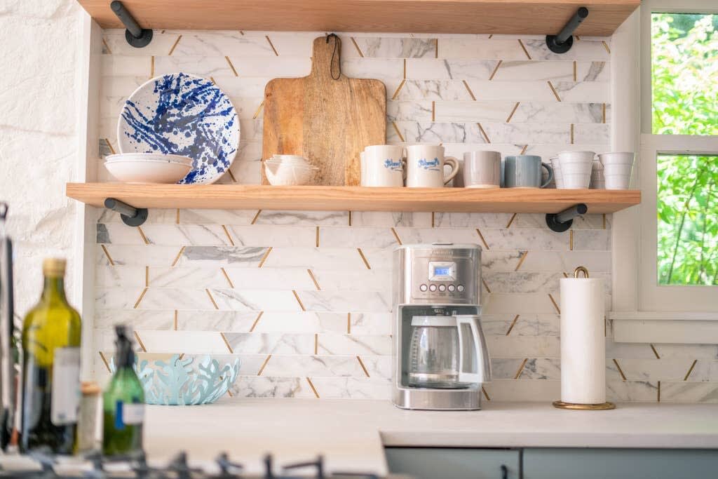 Close-up of a kitchen counter with a coffee maker, paper towel holder, and open wooden shelves displaying dishes and cutting boards against a white and gold patterned tile backsplash.