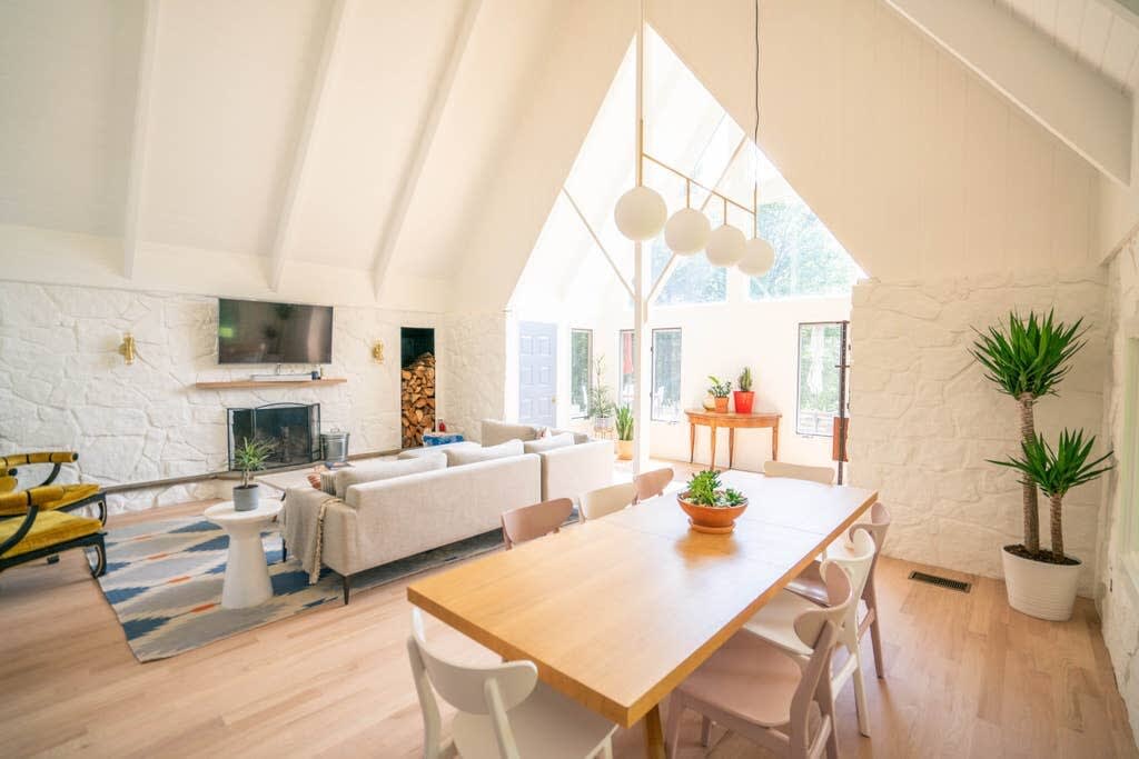 Spacious living and dining area with a high vaulted ceiling, light wood floors, and a large A-frame window, featuring a neutral sectional sofa, a long wooden dining table, and a white stone fireplace.