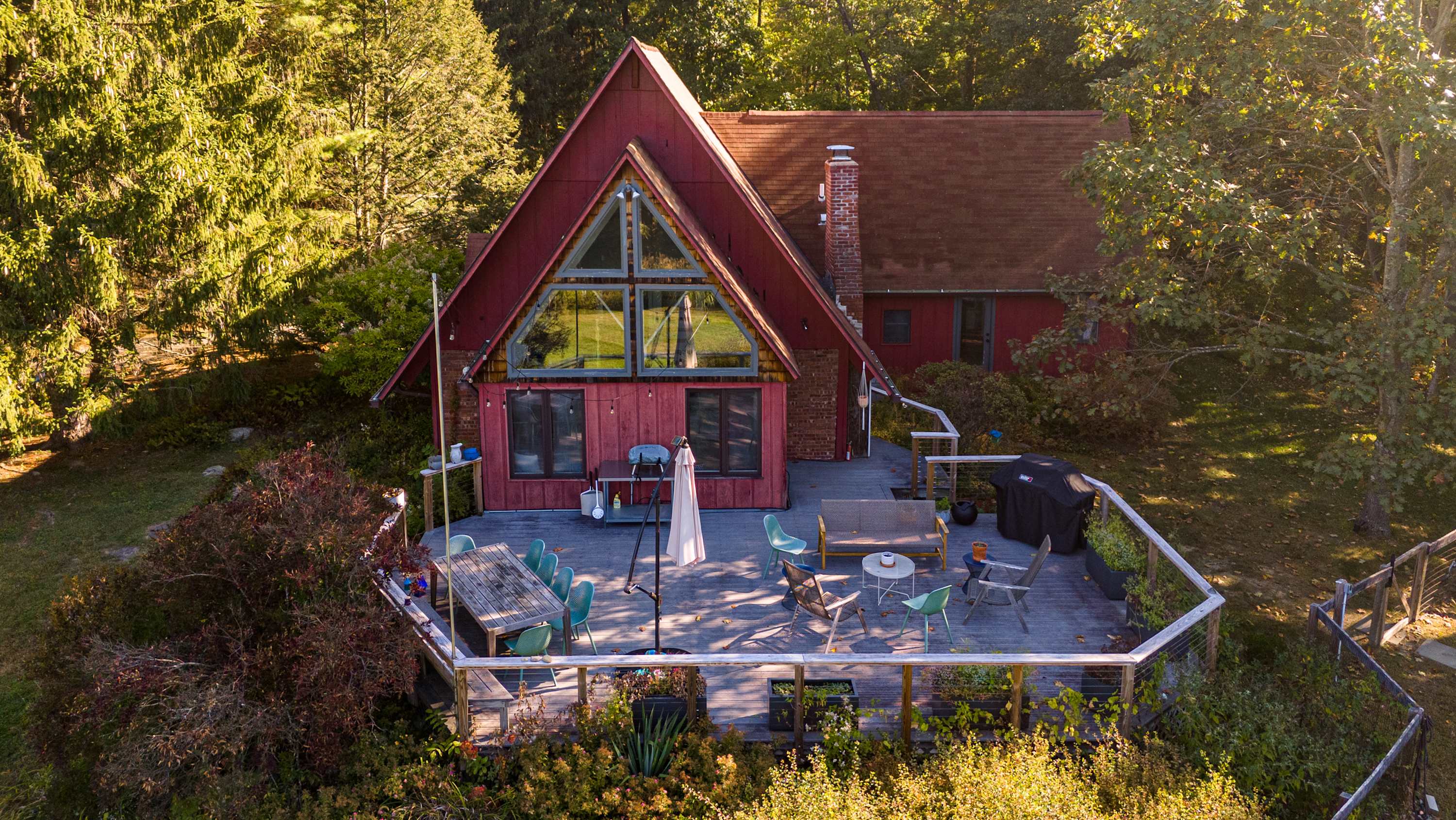 An aerial shot of the red A-frame house with its extensive wooden deck, surrounded by dense forest and autumnal trees. The deck features outdoor seating and dining areas, with a clear view of the natural landscape.