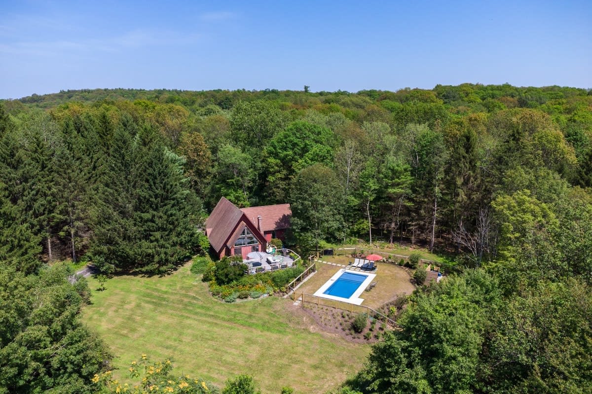 Aerial view of a red A-frame house nestled in a lush green forest, featuring a rectangular swimming pool with a white fence and a patio area with outdoor furniture. The property is surrounded by dense trees under a clear blue sky.