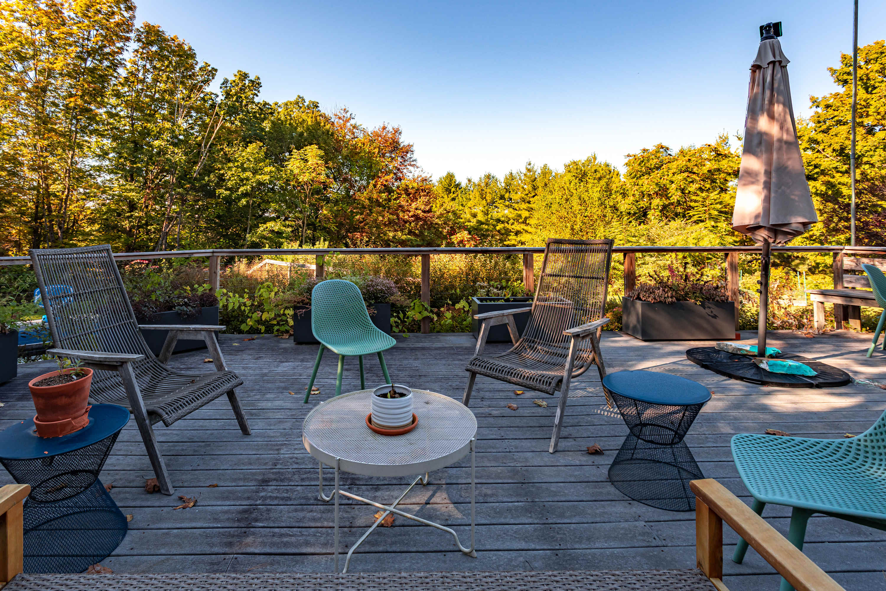A wooden deck furnished with various outdoor chairs and small tables, set against a backdrop of vibrant green and autumnal trees. A large closed umbrella stands in the background, suggesting a comfortable outdoor lounging area.