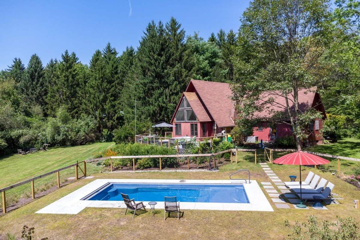 A high aerial view of the red A-frame house and its large wooden deck, situated amidst a sprawling green landscape and dense forest. A rectangular swimming pool with lounge chairs and a red umbrella is visible in the foreground, enclosed by a fence.