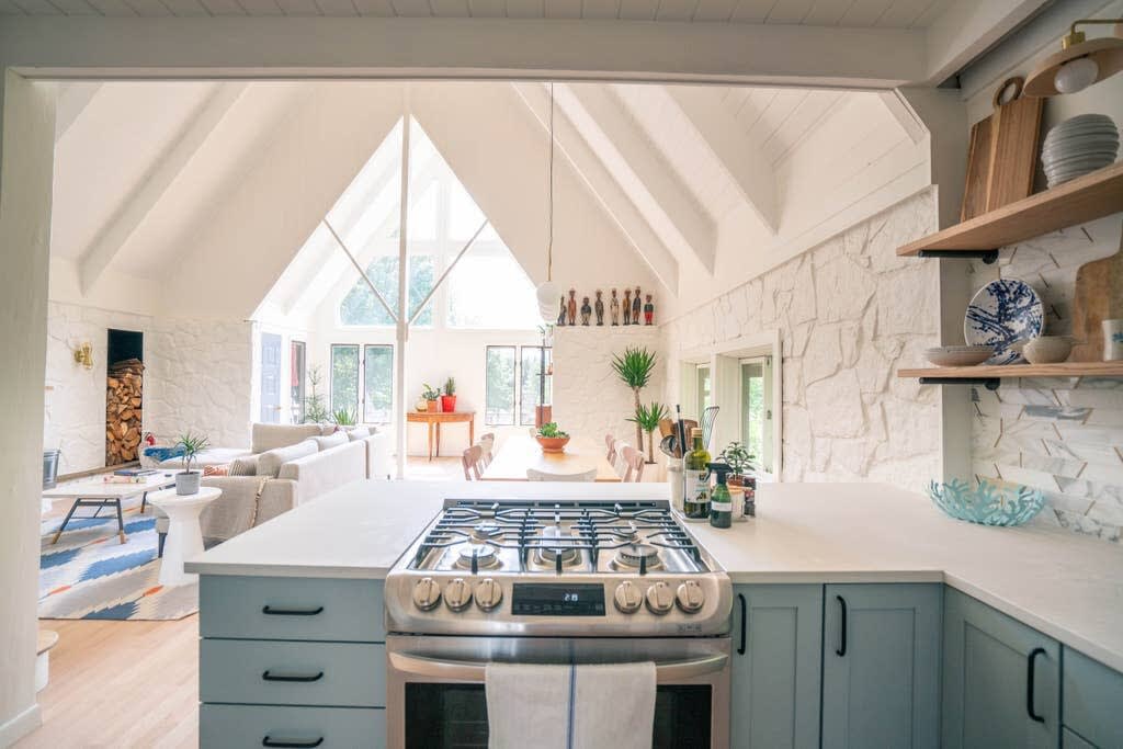 View from the kitchen into the open-concept living and dining area, showcasing a stainless steel gas range on a light countertop with light blue cabinets. The vaulted white ceiling extends through both spaces, with large A-frame windows visible in the distance.