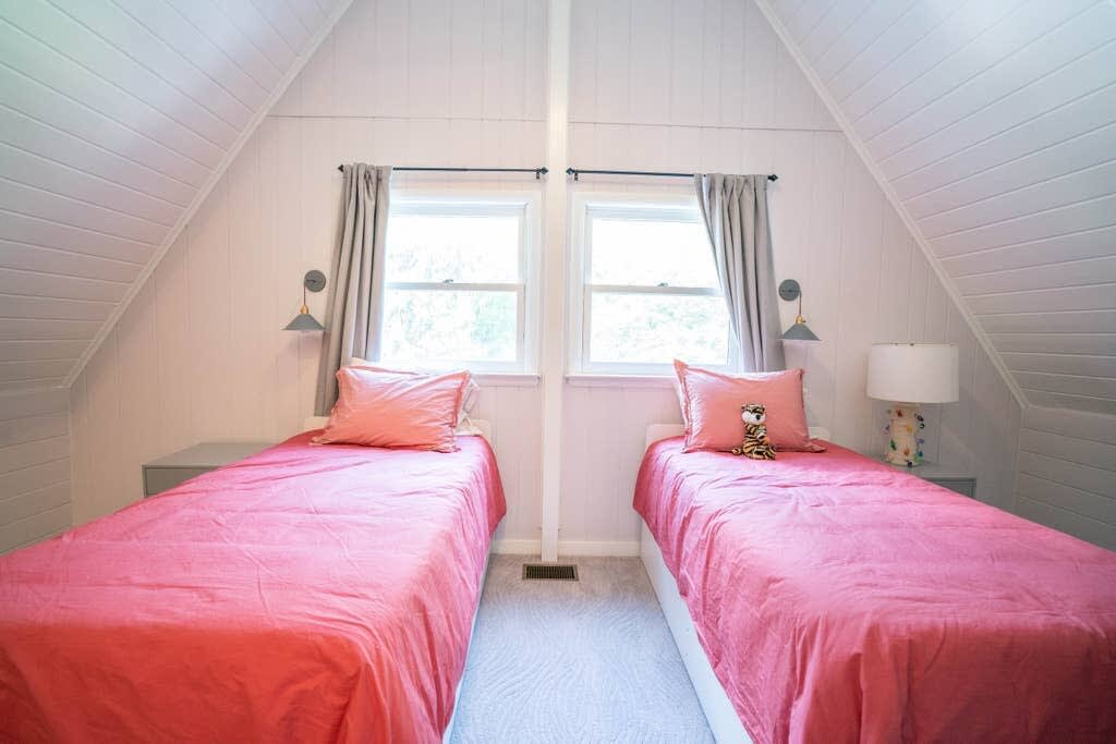 A cozy attic bedroom with two twin beds dressed in coral pink bedding, set against white paneled walls and a sloped ceiling. Each bed has a small side table and a wall-mounted sconce light.