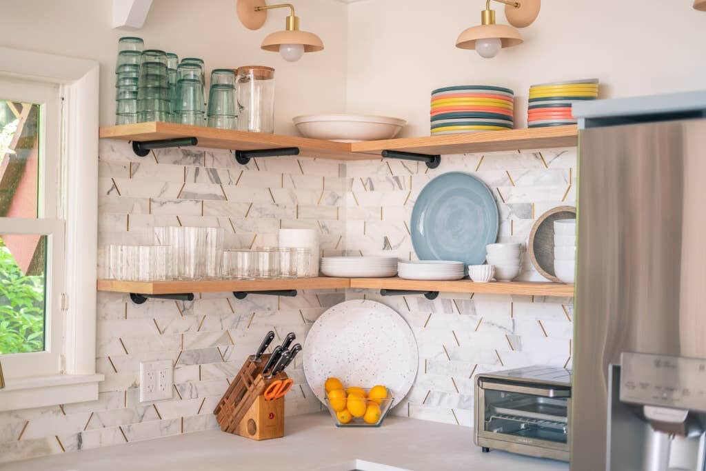 Detailed view of the kitchen's open wooden shelving against a white and gold geometric tile backsplash, displaying various glassware, dishes, and kitchen essentials. A stainless steel refrigerator and a toaster oven are visible on the white countertop.