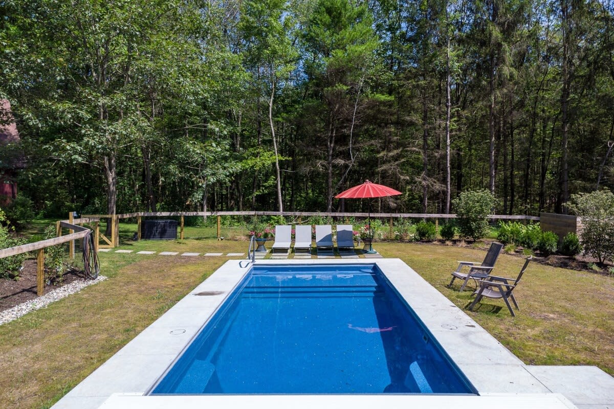 A rectangular inground swimming pool with a bright blue interior, surrounded by a white concrete patio and a grassy lawn. Several lounge chairs and a red umbrella are set up beside the pool, with a dense forest in the background.