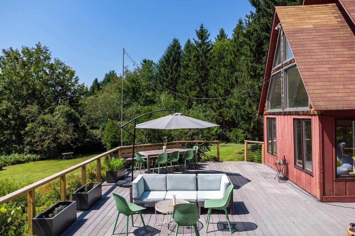 A wide view of the expansive wooden deck attached to the red A-frame house, featuring a modern outdoor sofa, green chairs, and a dining set under a large umbrella. The deck overlooks a vast green lawn and dense forest.