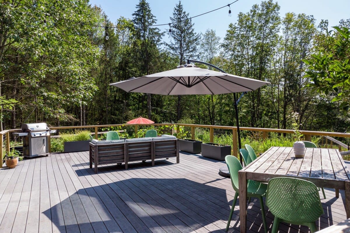 Spacious outdoor deck with gray composite decking, featuring a large patio umbrella, a gas grill, a wooden dining table with green chairs, and a lounge seating area, surrounded by lush trees.