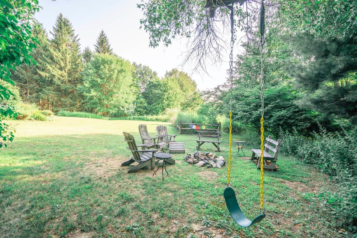 A rustic outdoor gathering area on a grassy lawn, centered around a stone fire pit. It is furnished with wooden Adirondack chairs, benches, and a green swing hanging from a tree, surrounded by dense forest.