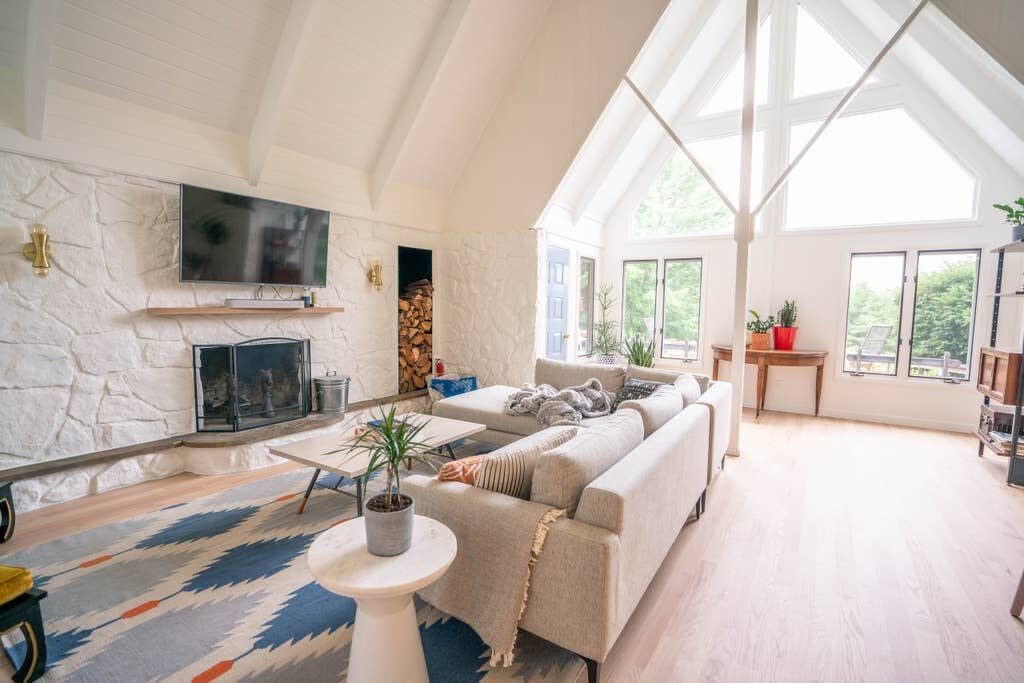 Spacious living room with a vaulted white ceiling and exposed beams, featuring a large L-shaped sectional sofa on a colorful geometric rug. A white stone fireplace with a wooden mantel and a mounted TV anchors one wall, with large windows offering views of the outdoors.