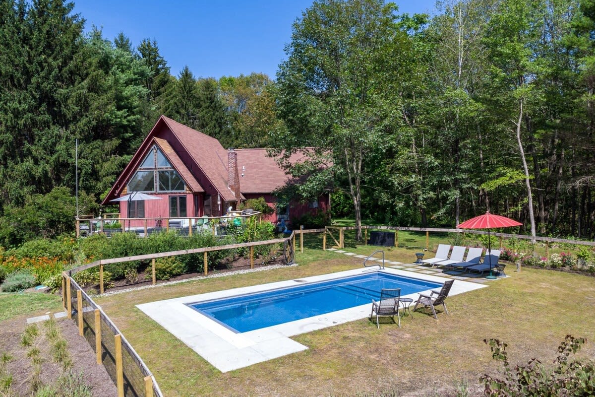 An aerial view of a red A-frame house nestled among lush trees, featuring a large inground swimming pool with a white surround in the foreground. A low wooden fence encloses the pool area, which includes lounge chairs and an umbrella.