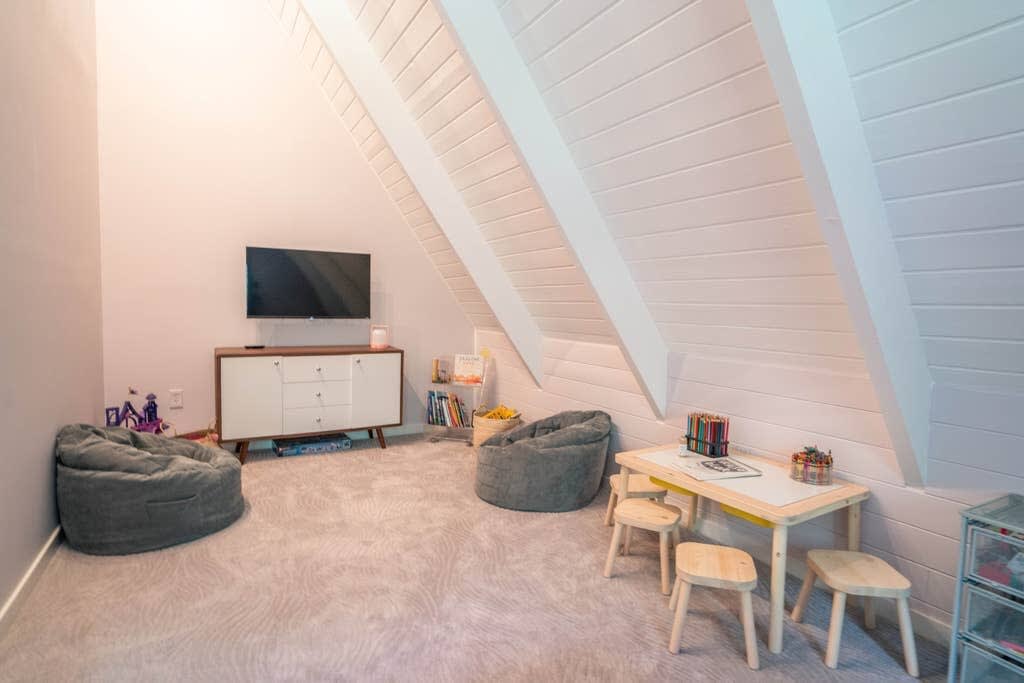 A bright play area with a large TV on a white credenza, two grey bean bag chairs, and a small wooden table with stools for children. The room features a sloped ceiling with white paneling and a light-colored patterned carpet.