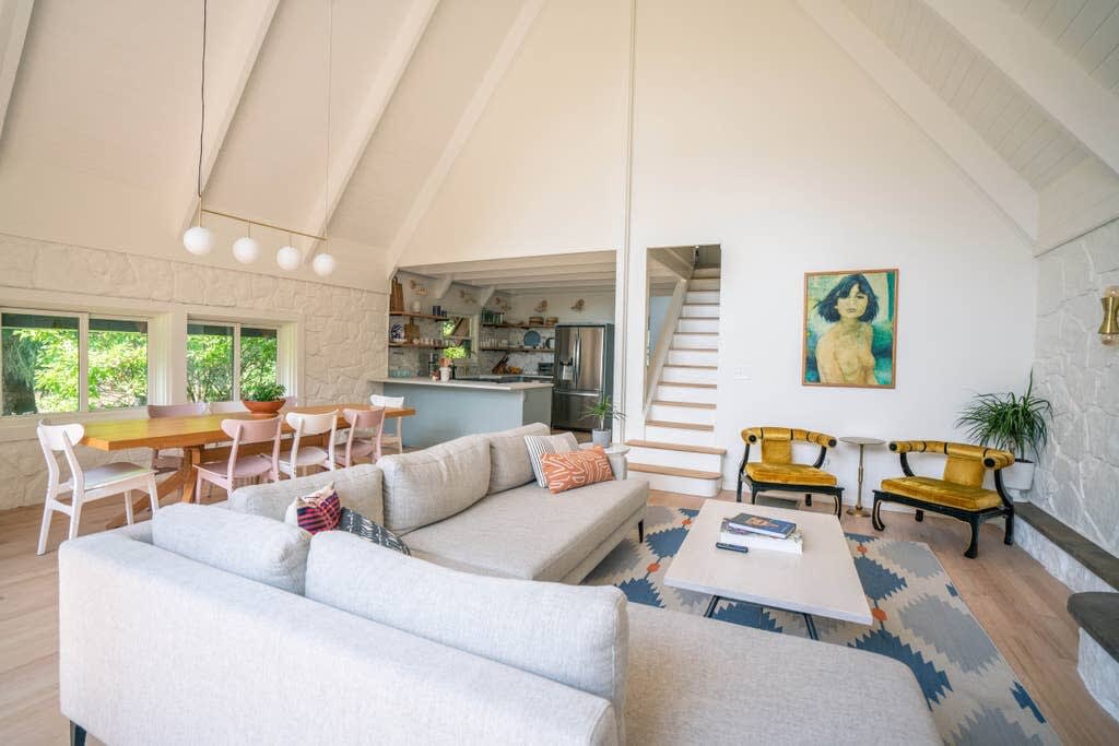 Open-concept living and dining area with a high vaulted ceiling and light hardwood floors, featuring a large sectional sofa and a wooden dining table with mixed chairs. A modern chandelier hangs over the dining area, and a colorful abstract painting adorns the wall.