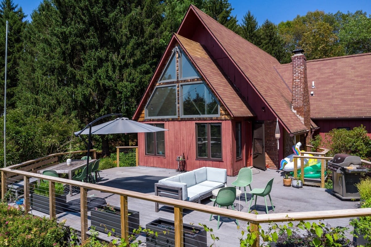 An A-frame house with a red exterior and large triangular windows, surrounded by lush green trees. A spacious wooden deck with outdoor seating, a dining area, and a grill extends from the house.