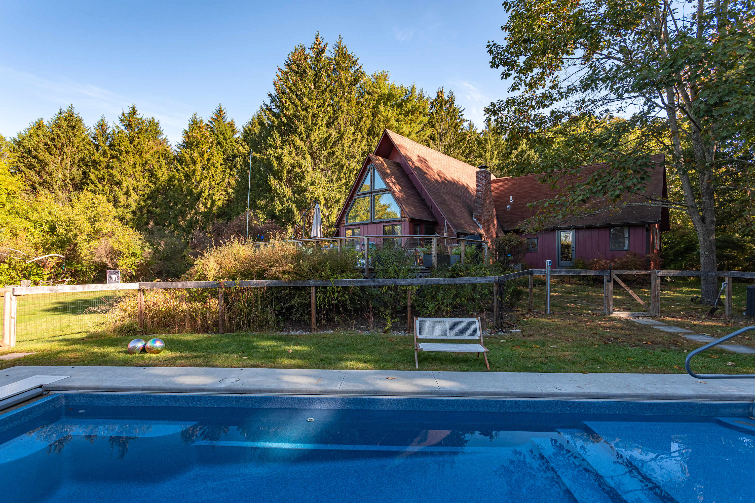 View of the red A-frame house from the pool area, showing a large deck with a railing, surrounded by lush greenery and tall evergreen trees under a clear blue sky.