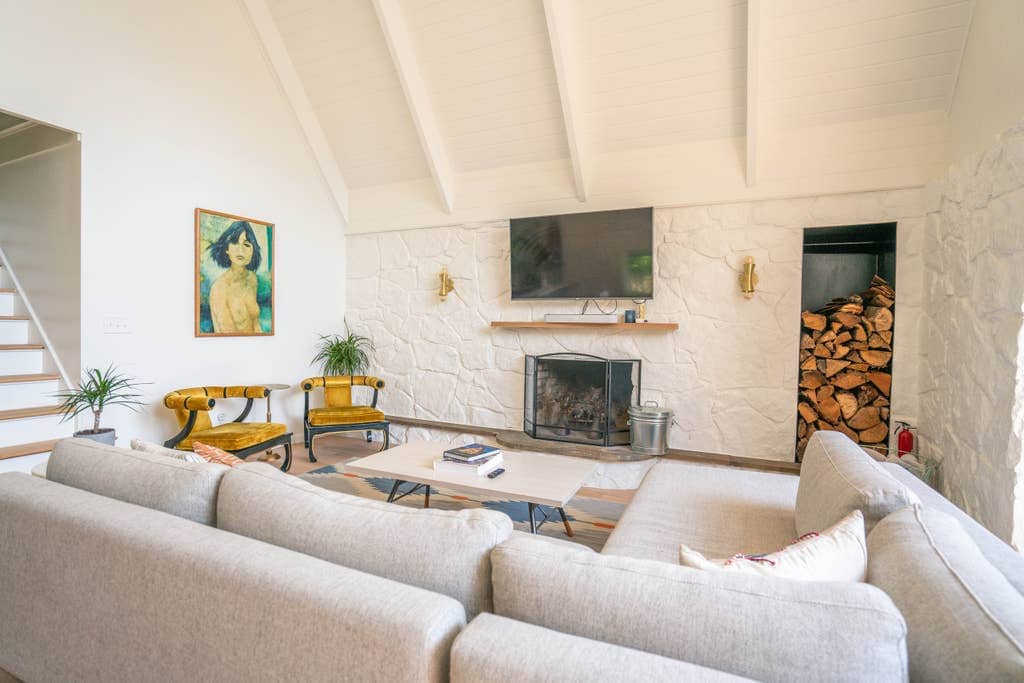 Cozy living room area with a large neutral sectional sofa, a white stone fireplace with a wooden mantel, a wall niche filled with firewood, and a flat-screen TV mounted above.
