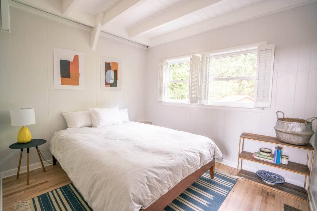 A bright bedroom featuring a queen bed with white bedding, light wood floors, and white paneled walls. Two abstract art prints hang above the bed, and a yellow lamp sits on a small side table.