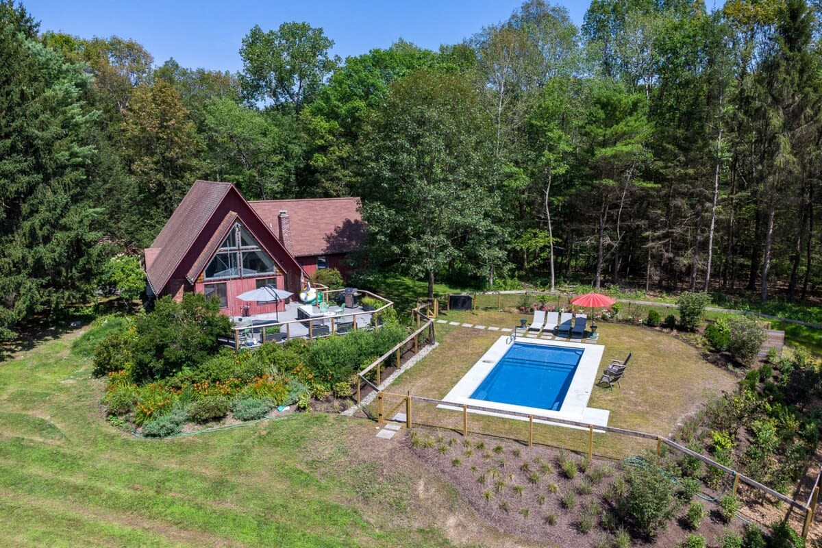 An aerial view showcasing the red A-frame house with its large deck, nestled among lush green trees. A rectangular swimming pool with a surrounding lawn and lounge chairs is visible in the foreground, connected by a path.