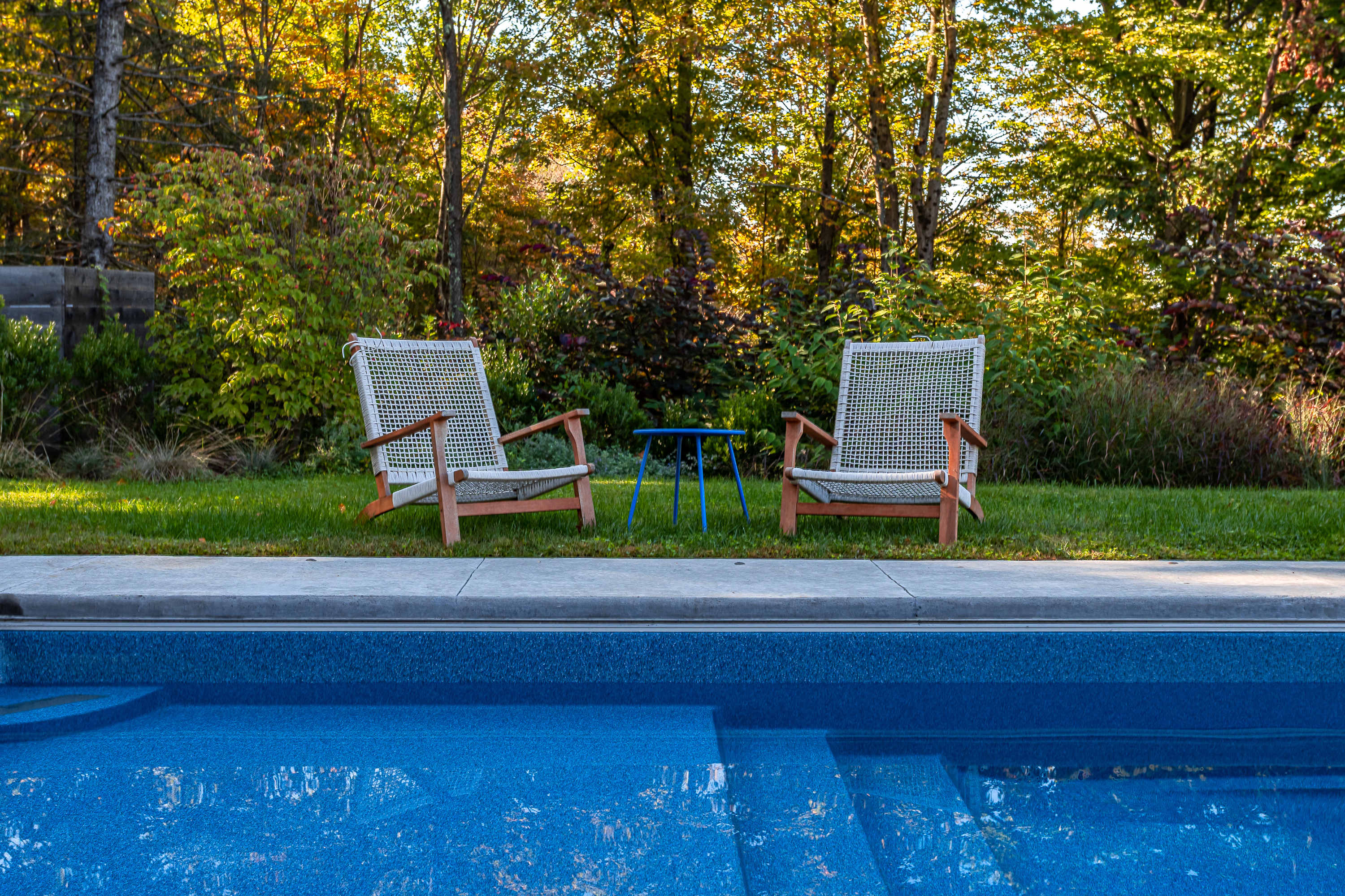 Two modern lounge chairs with wooden frames and woven seats positioned on a green lawn next to the edge of a blue inground swimming pool, surrounded by lush trees.
