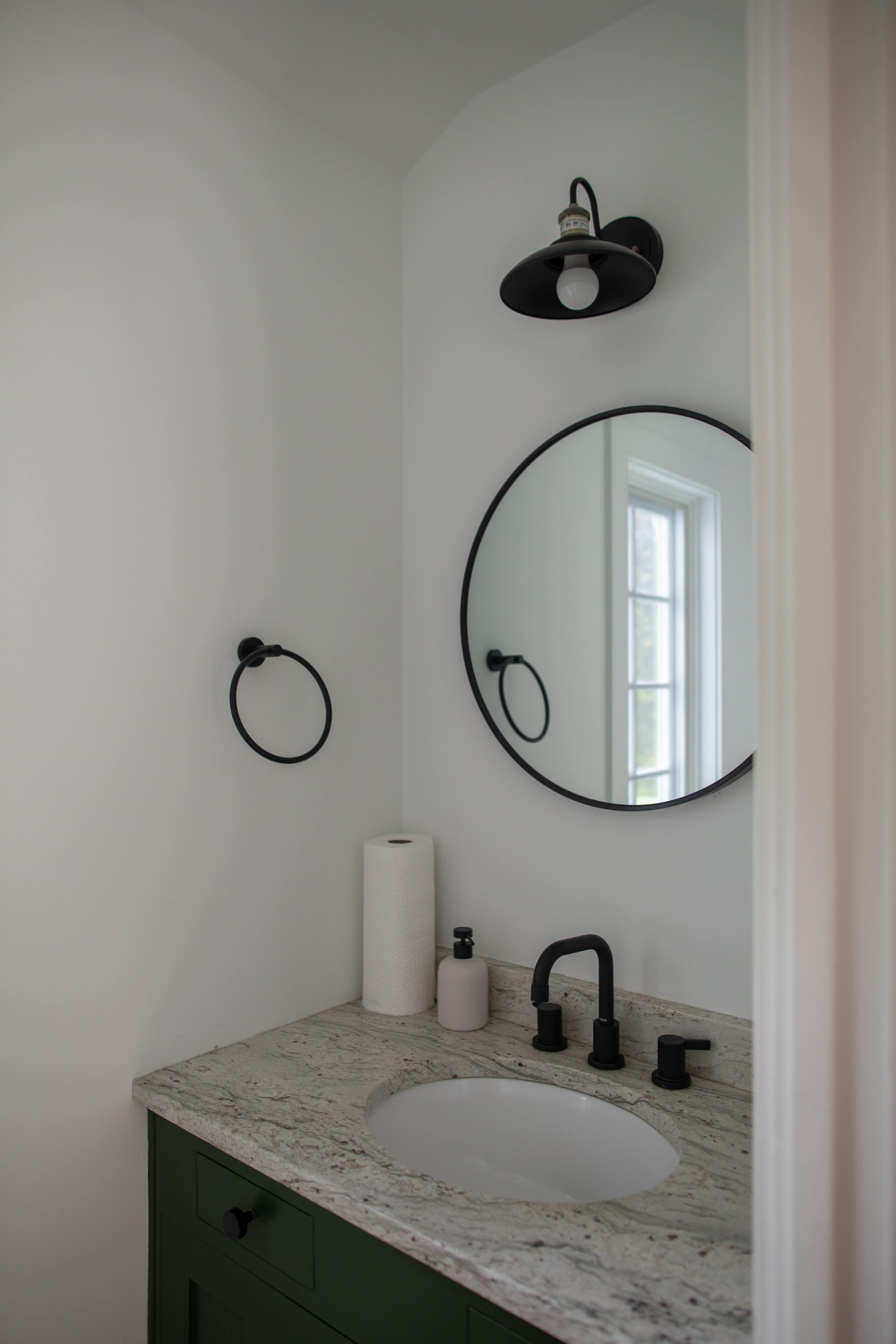 A modern powder room featuring a dark green vanity with a granite countertop and an oval sink, complemented by a black circular mirror and black fixtures. A black industrial-style sconce provides lighting above the mirror.