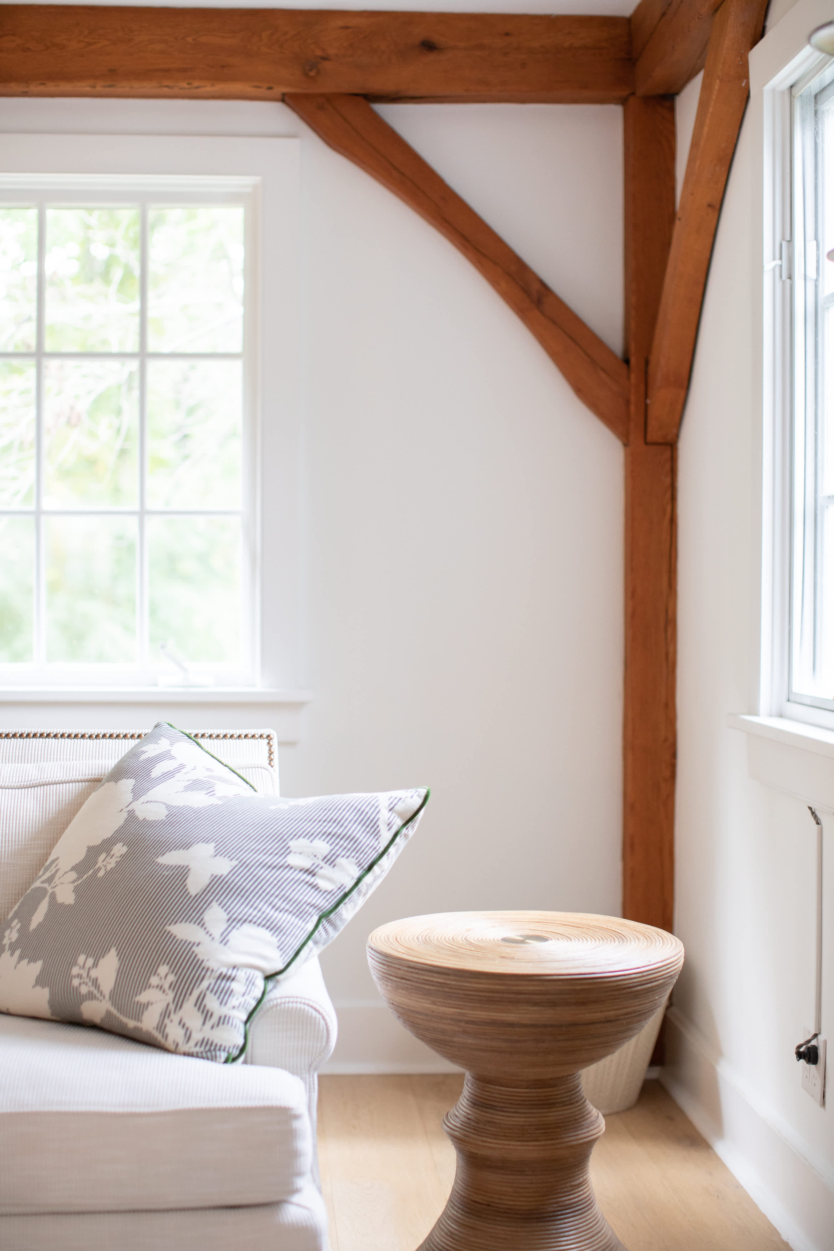 A close-up of a living room corner, highlighting a cream armchair with a patterned pillow and a unique wooden side table with a ribbed design. A large window offers a view of lush greenery, and a rustic wooden beam is visible in the background.