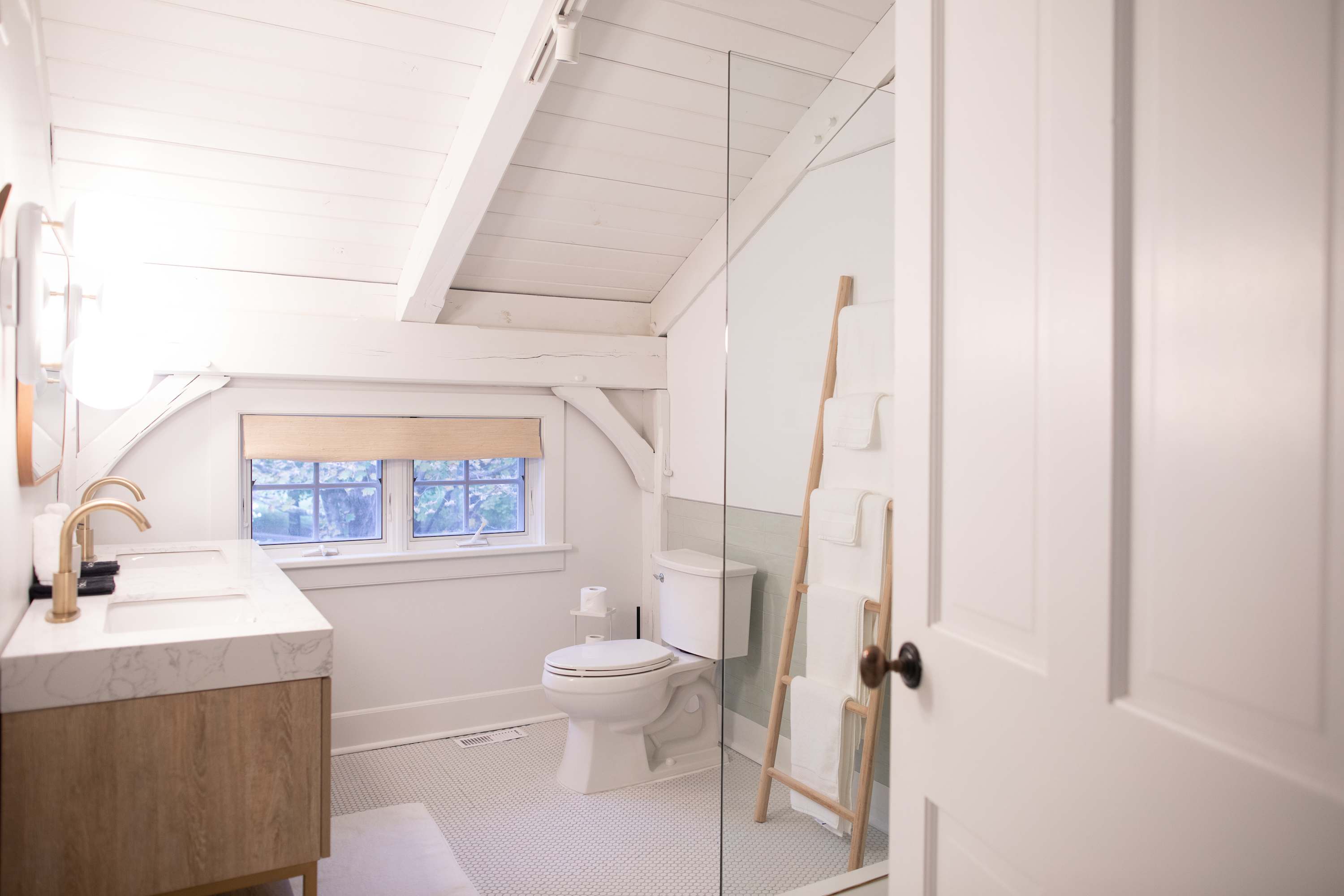 A bright bathroom with white shiplap vaulted ceilings and small hexagonal tile flooring, featuring a modern wooden vanity with double sinks and gold fixtures. A large glass shower enclosure is visible, along with a toilet and a wooden ladder towel rack.