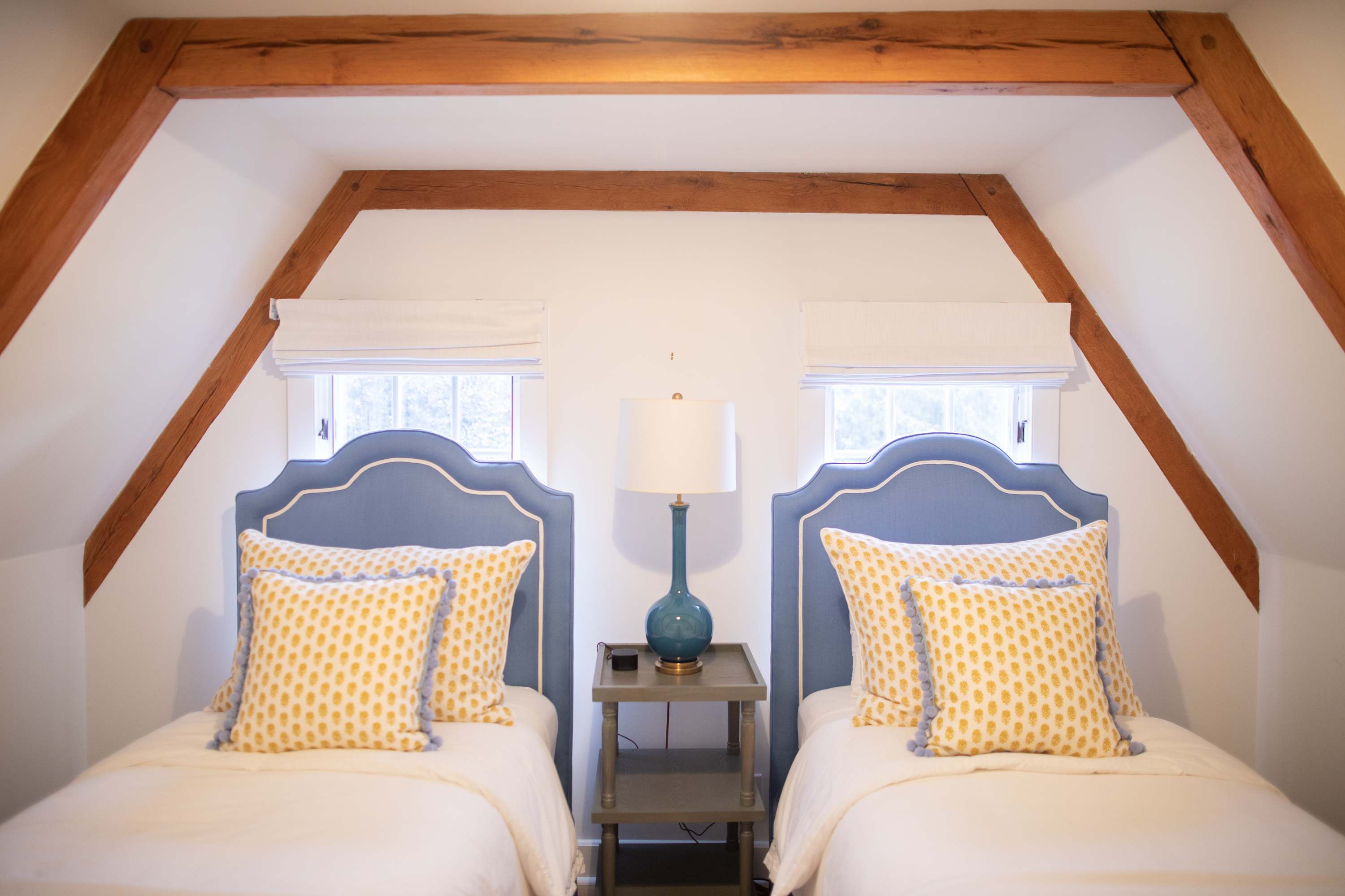 Cozy attic bedroom featuring two twin beds with blue upholstered headboards, yellow patterned pillows, and a shared nightstand with a teal lamp, all beneath exposed wooden beams.