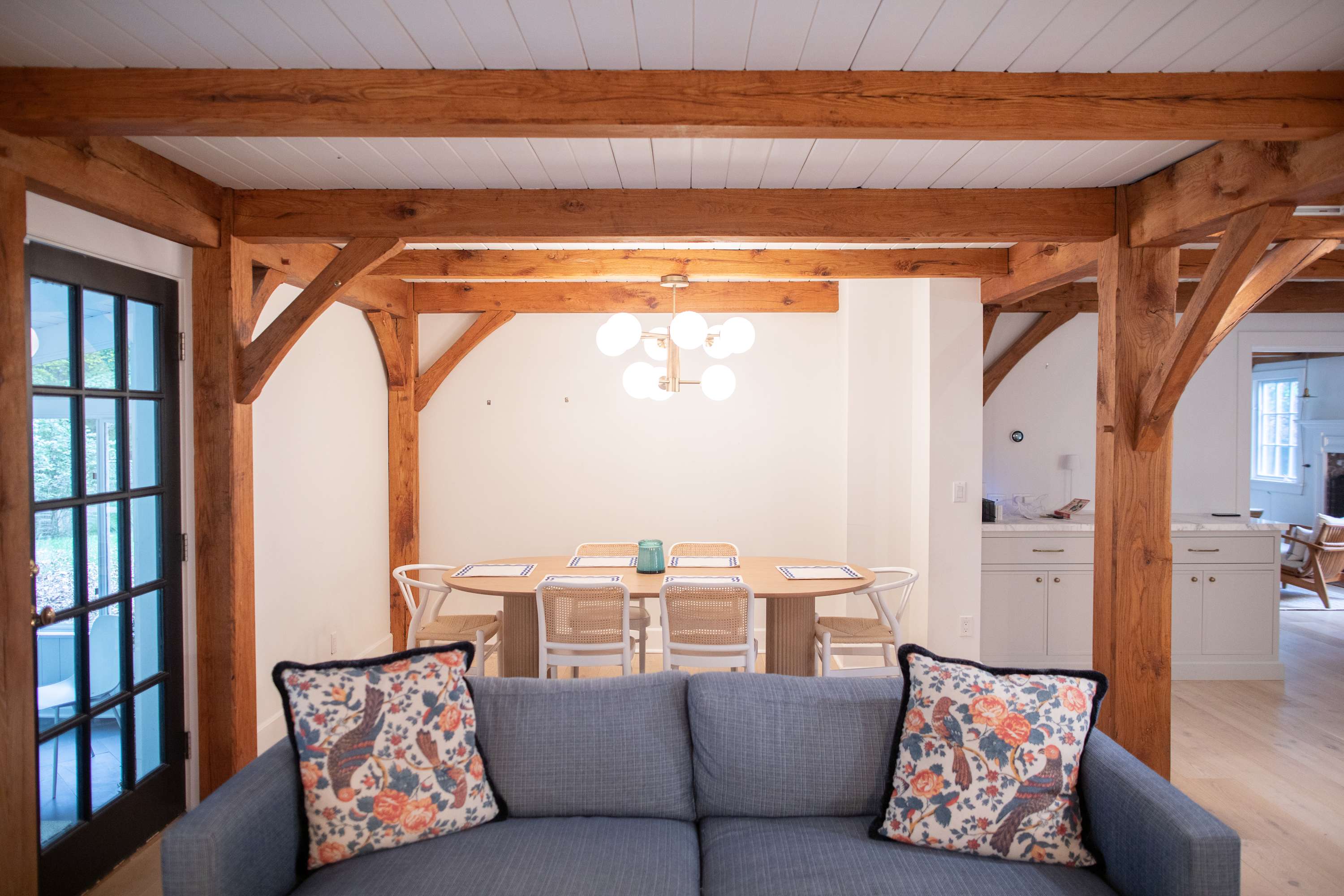 A bright dining area with light hardwood floors and exposed wooden beams, featuring an oval wooden dining table surrounded by modern chairs. A contemporary chandelier hangs above, and a blue sofa with patterned pillows is visible in the foreground.