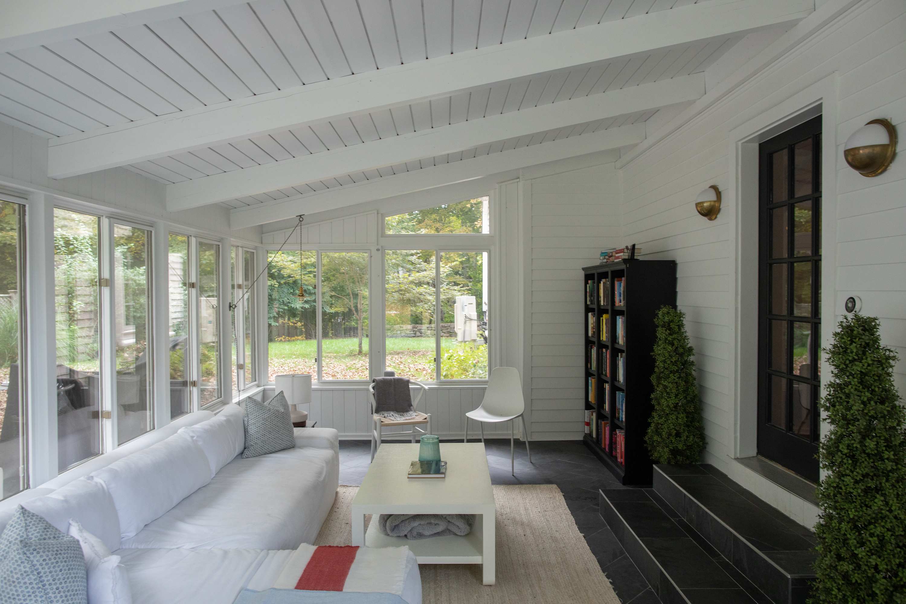 Bright sunroom with a white slipcovered sofa, a white coffee table, and large windows offering views of the outdoors, featuring a vaulted white ceiling with exposed beams and dark tiled flooring.
