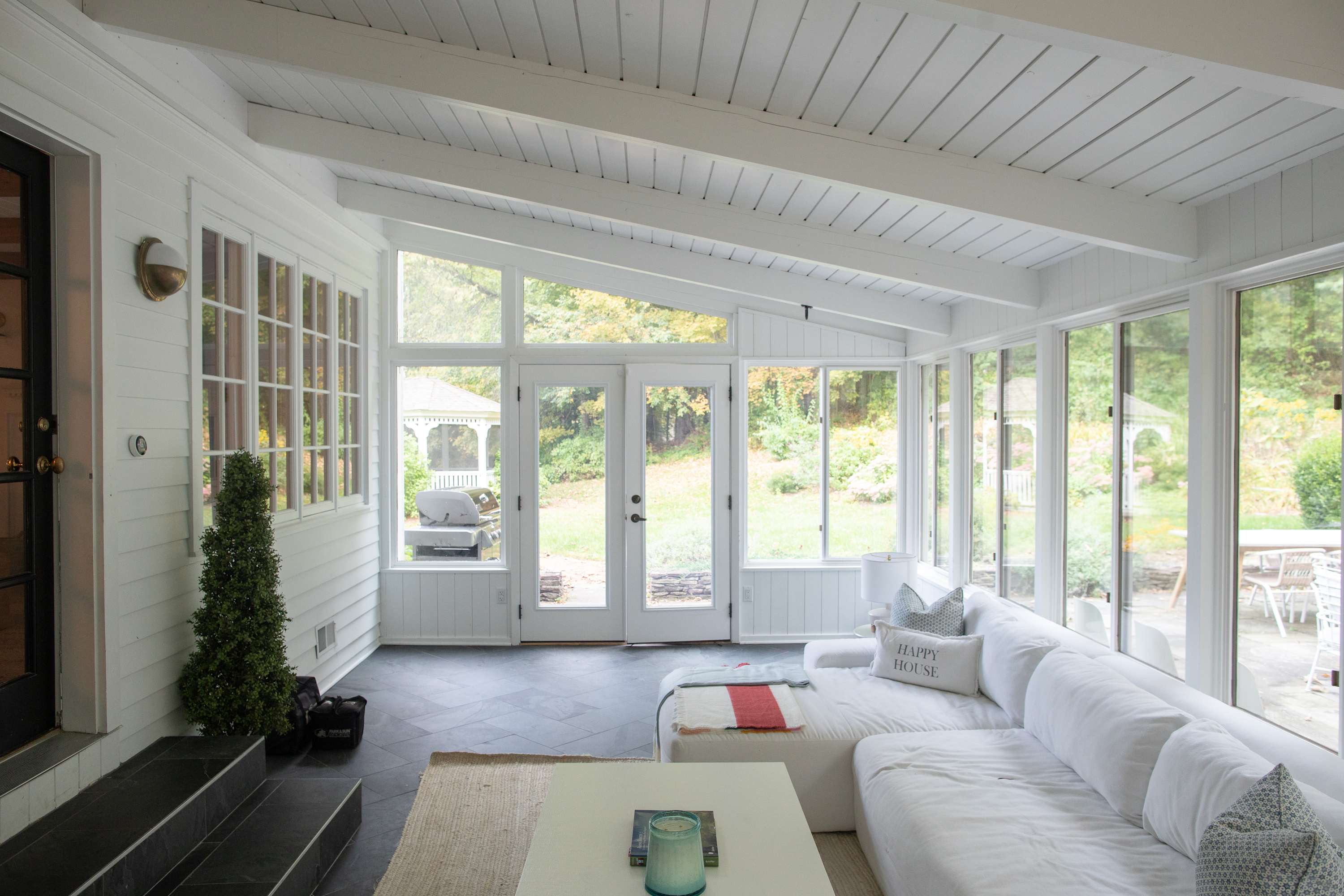 Bright sunroom with white paneled walls and ceiling, featuring large windows and glass doors that open to the outdoor yard. A large white sectional sofa provides ample seating, creating a comfortable space to enjoy the views.