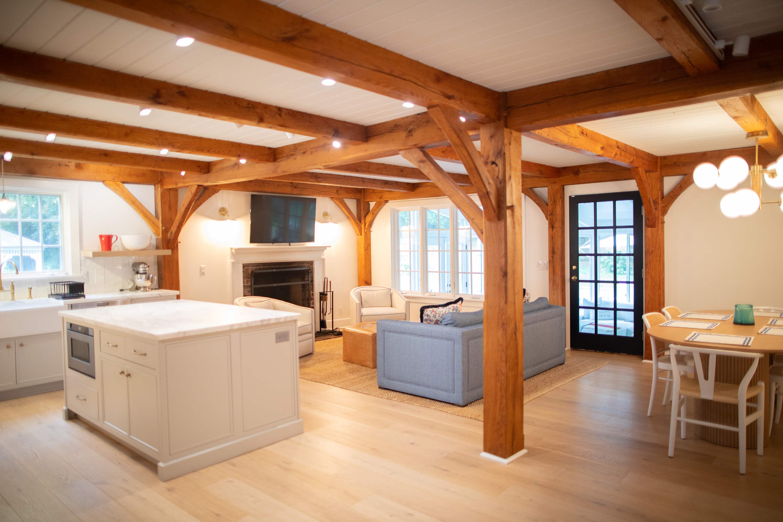 An expansive open-concept living space with light hardwood floors and exposed wooden beams on the ceiling. The area includes a modern kitchen island, a cozy living room with a fireplace, and a dining area with a round table, all bathed in natural light.