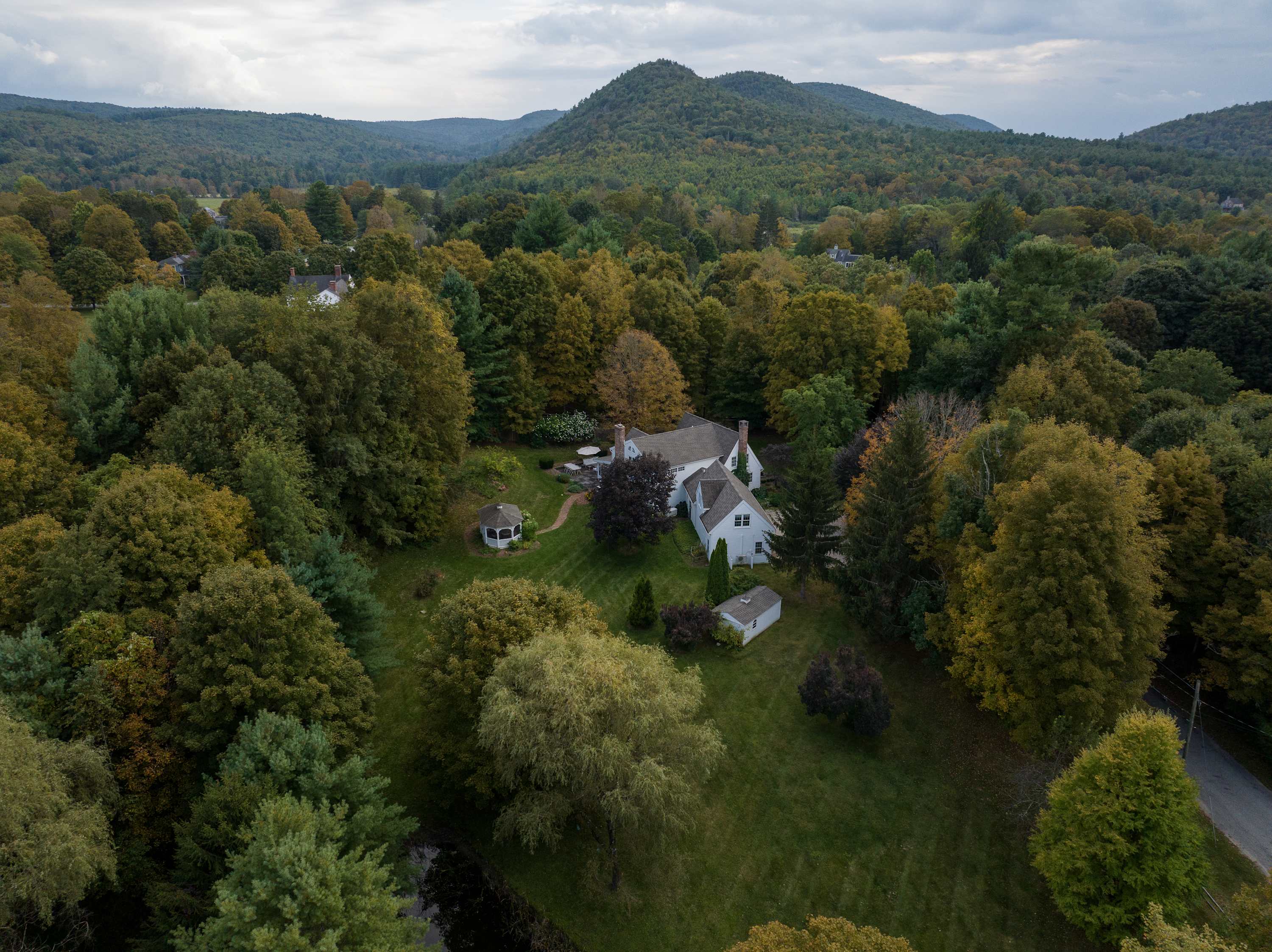 An expansive aerial view showcases the white house nestled among a dense forest of green and autumn-colored trees, with a distant mountain range visible under a cloudy sky. The property includes a gazebo and a large grassy yard.