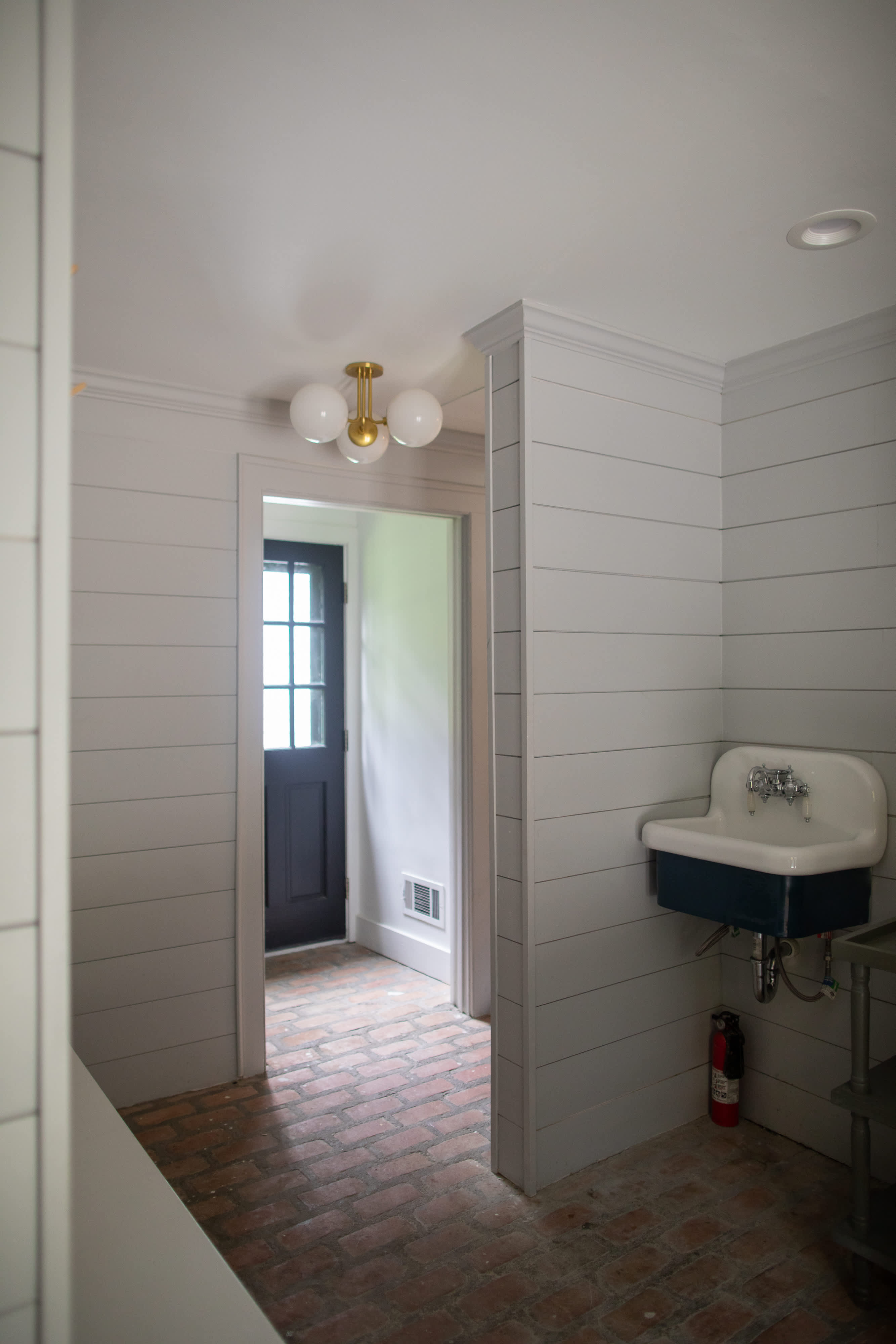 A rustic mudroom featuring light gray shiplap walls, a dark blue wall-mounted utility sink with exposed plumbing, and a red brick floor. A dark door with glass panes leads to the outside, illuminated by a modern brass and globe light fixture.