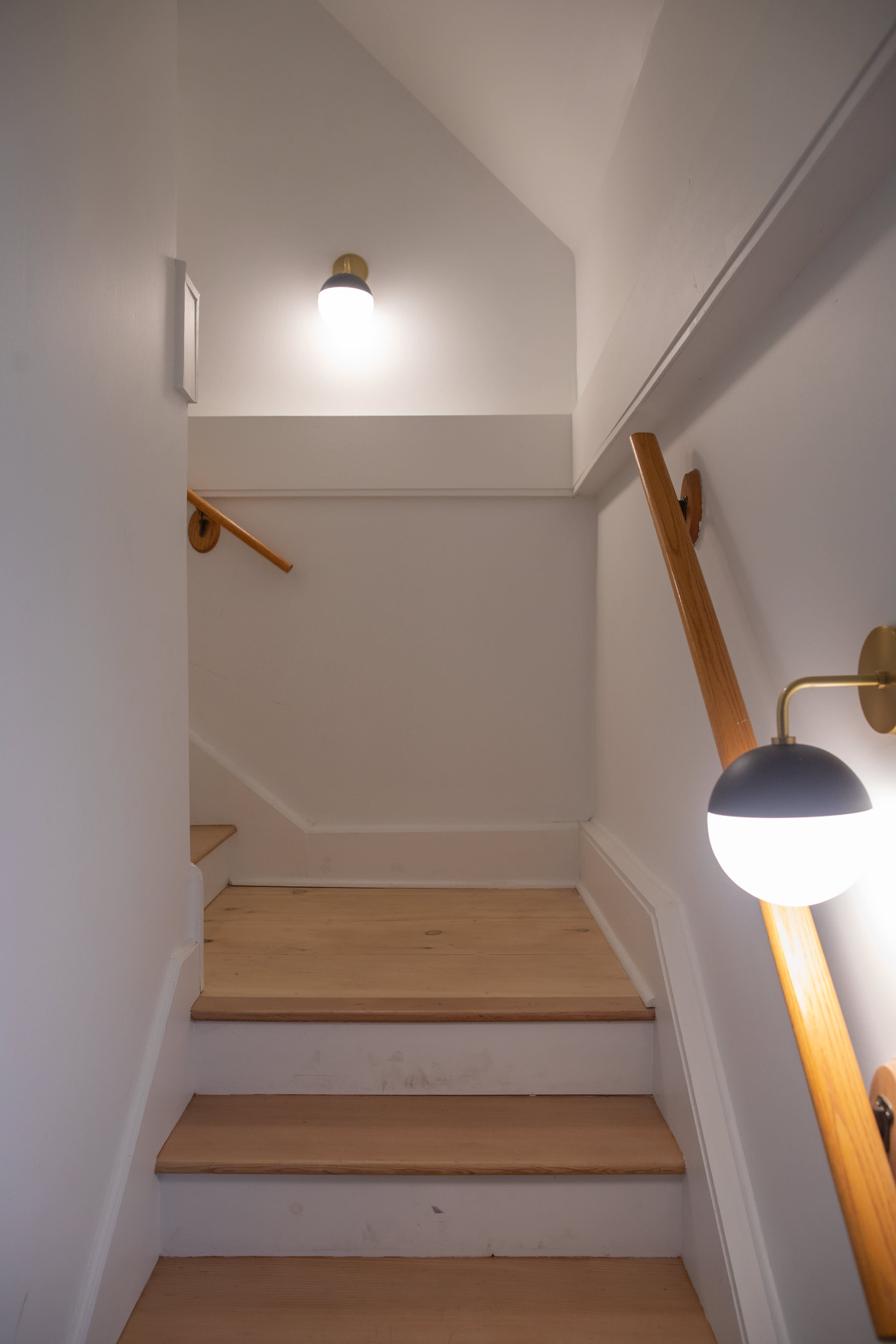 Well-lit wooden staircase with white risers and light wood treads, featuring modern wall sconces and wooden handrails, leading up to an upper level.