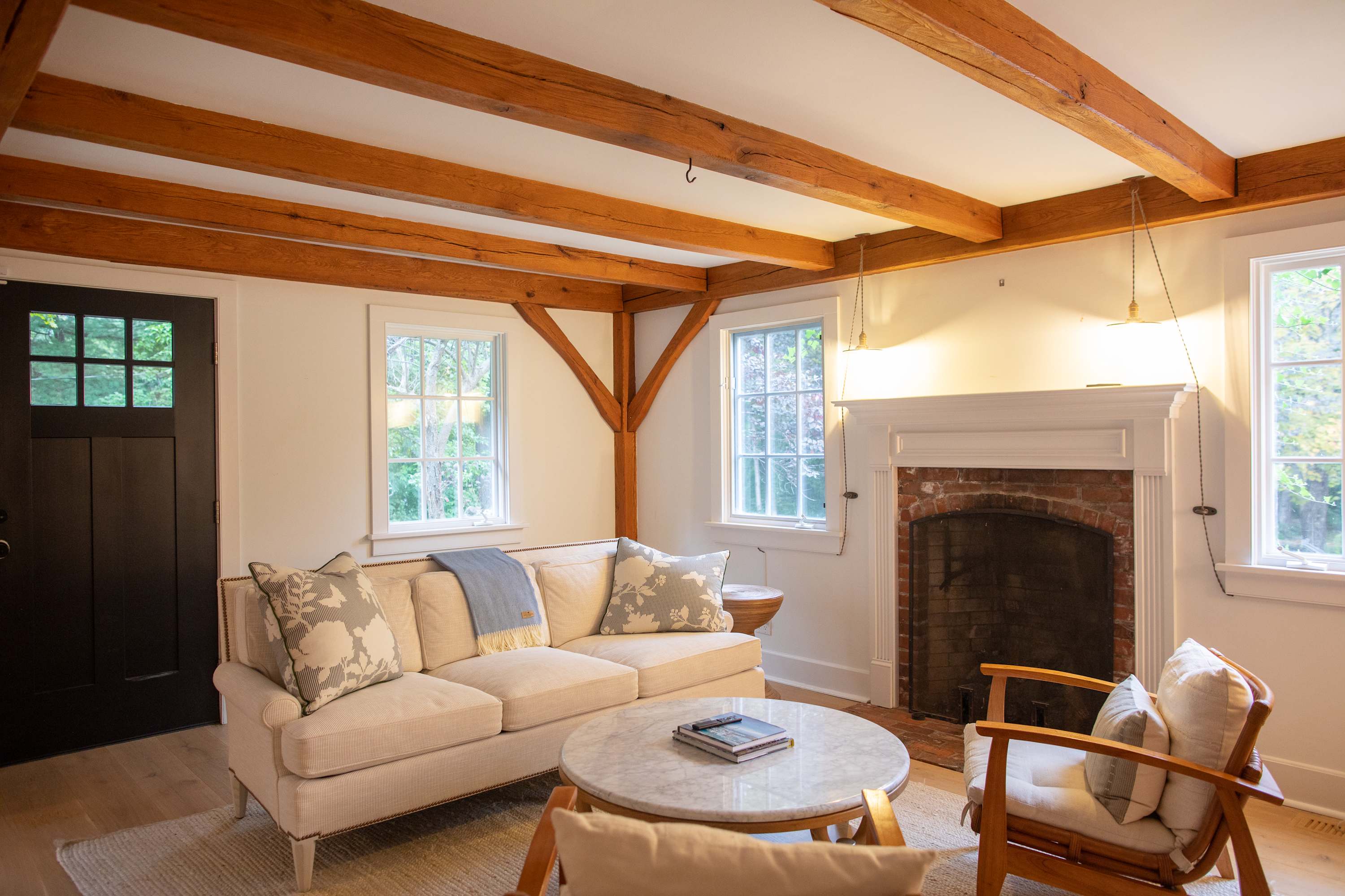 A warm living room with light hardwood floors and prominent exposed wooden beams, centered around a cream sofa with decorative pillows. A traditional brick fireplace with a white mantel anchors one wall, complemented by a round marble coffee table and two wooden armchairs.