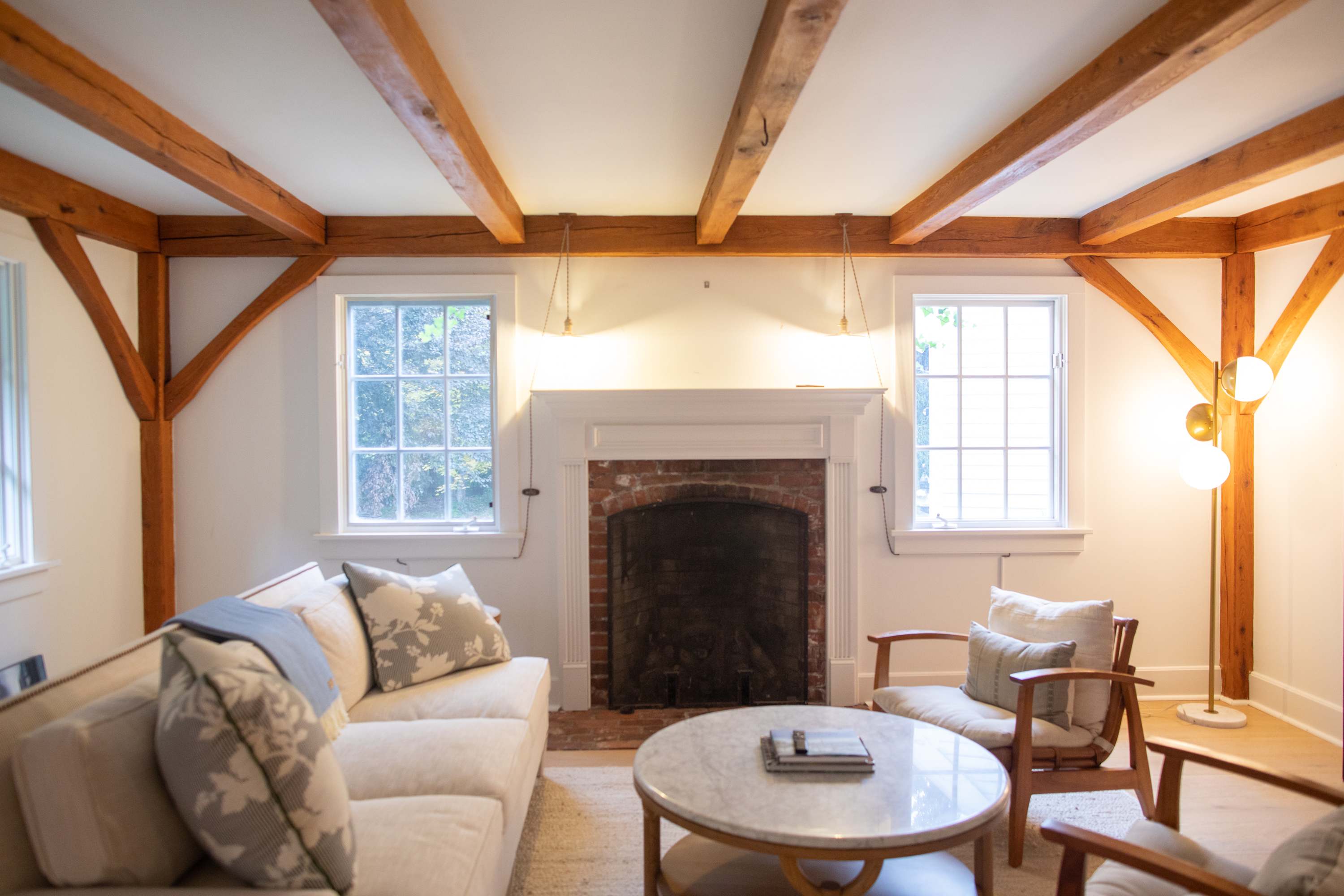 Cozy living room with exposed wooden beams on the white ceiling, a brick fireplace with a white mantel, and light-colored furniture including a sofa and armchairs arranged around a marble-topped coffee table. Two windows flank the fireplace, letting in natural light.