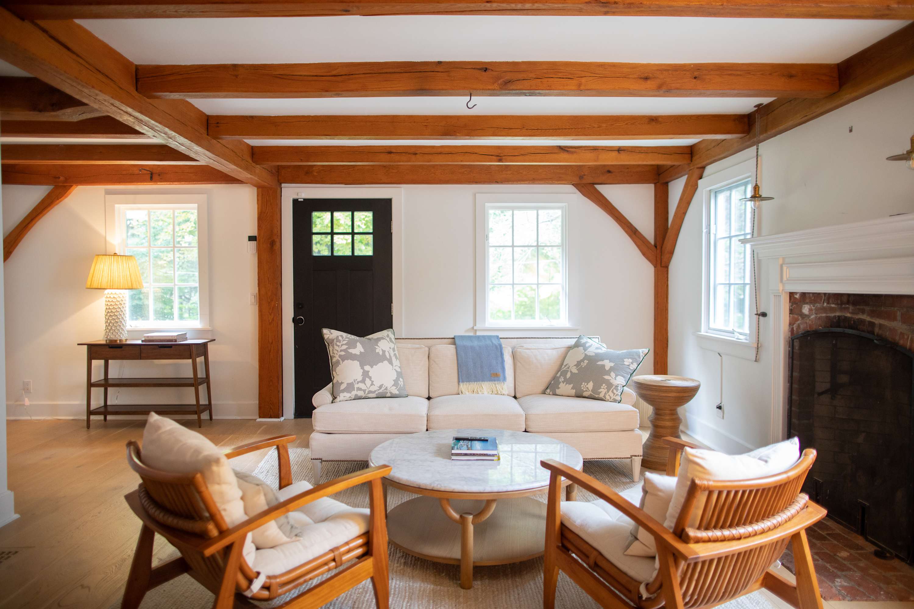 A cozy living room featuring light hardwood floors, exposed wooden ceiling beams, and a cream-colored sofa adorned with patterned pillows. Two wooden armchairs flank a round marble coffee table, all set against white walls with multiple windows.