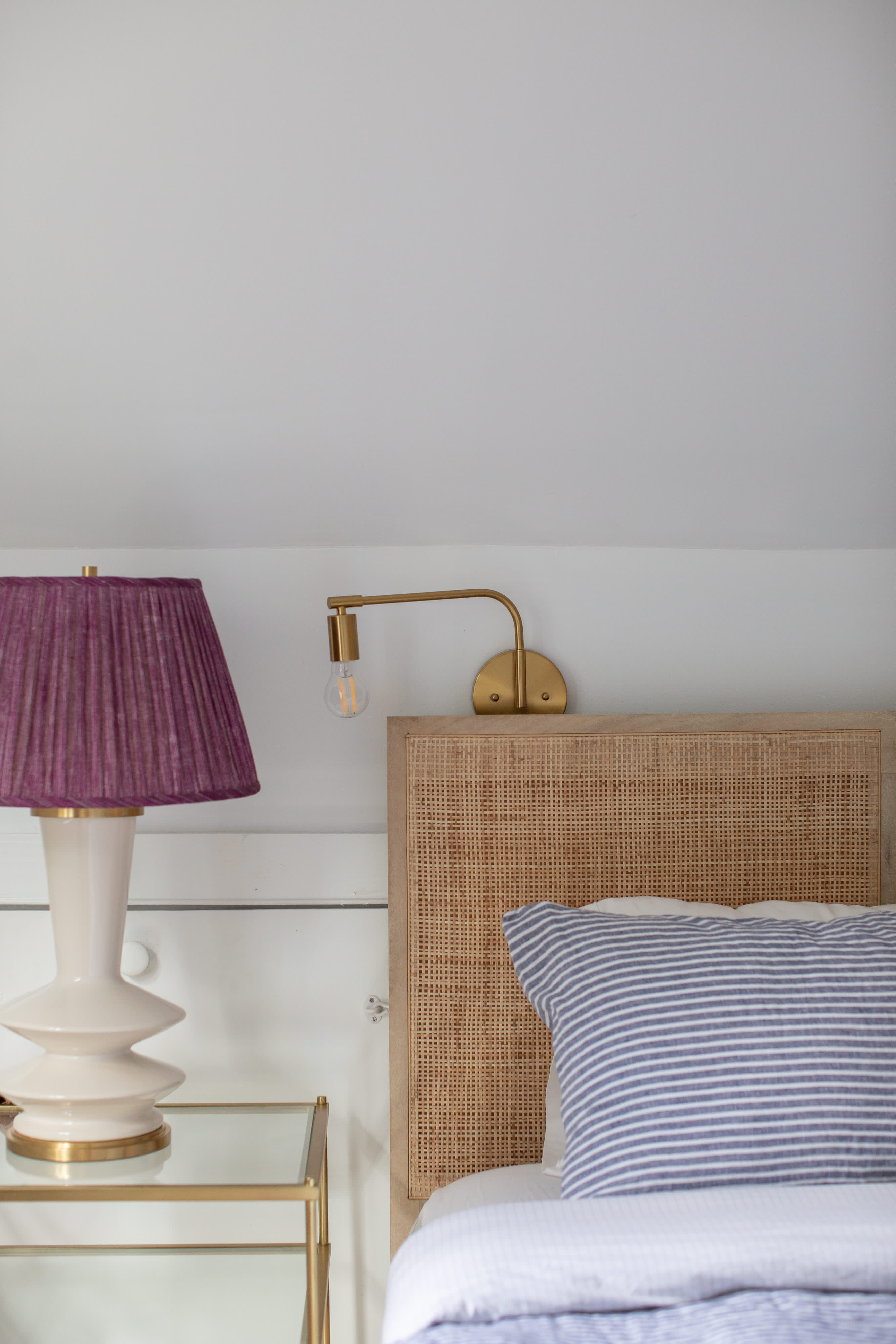 Close-up view of a bedroom featuring a natural rattan headboard, striped pillows, a gold wall sconce, and a white and purple pleated lamp on a glass and gold nightstand.
