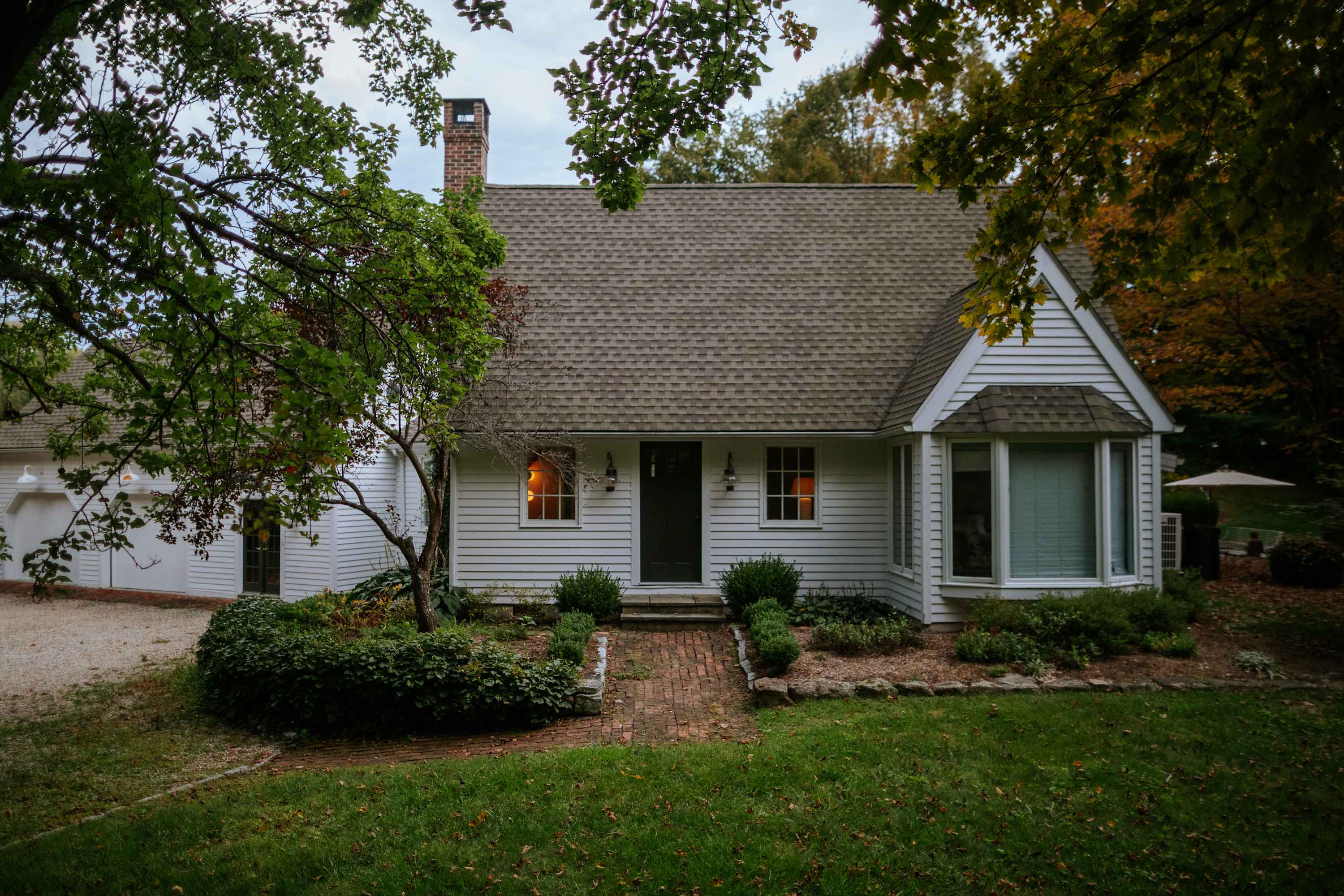 The front facade of the white clapboard house features a dark front door flanked by windows, a bay window on the right, and a brick pathway leading to the entrance. Lush green landscaping with shrubs and small trees frames the house.