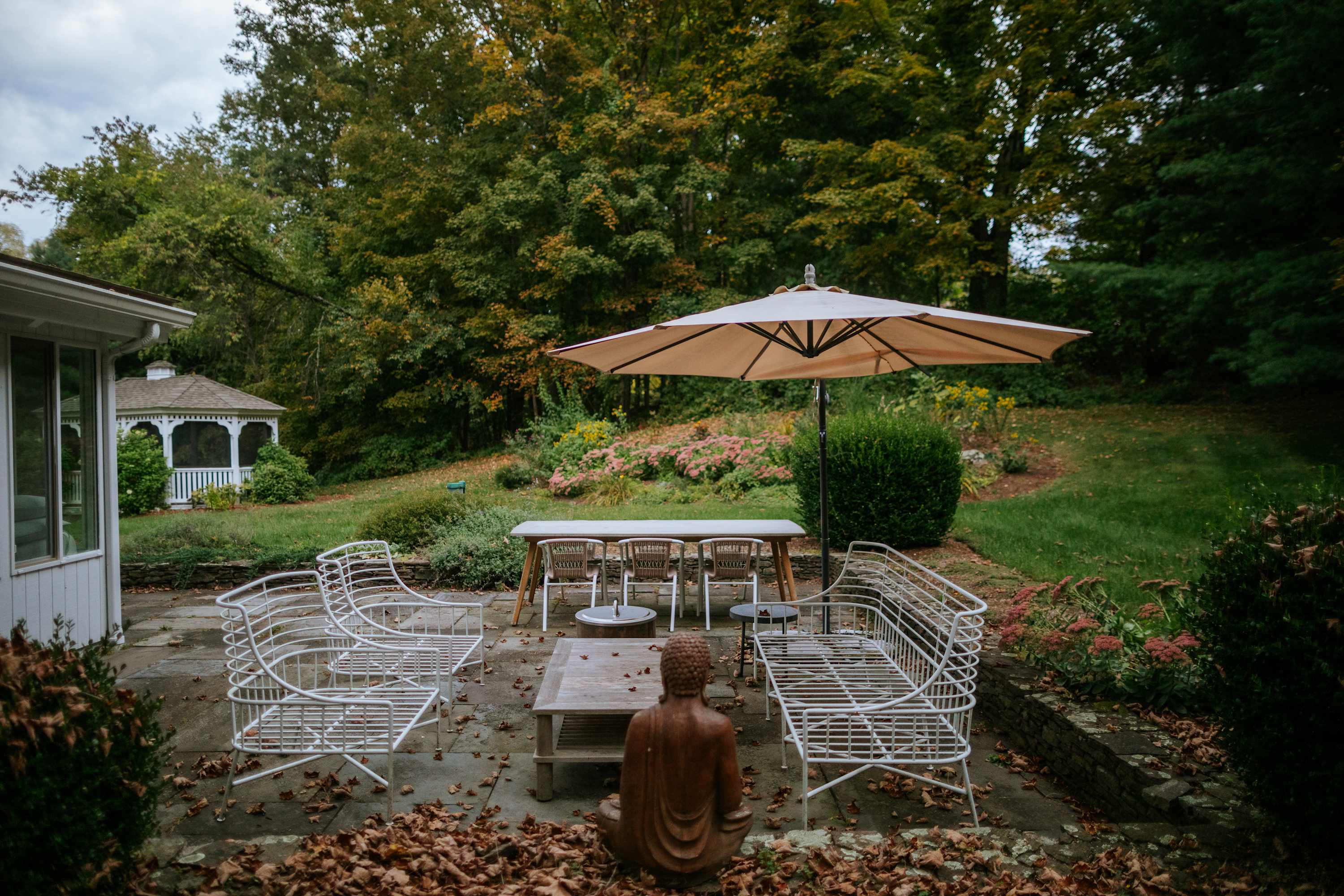 A spacious outdoor patio with stone pavers, featuring elegant white metal seating, a dining table with chairs, and a serene Buddha statue. Autumn leaves are scattered across the ground, surrounded by lush greenery and mature trees.