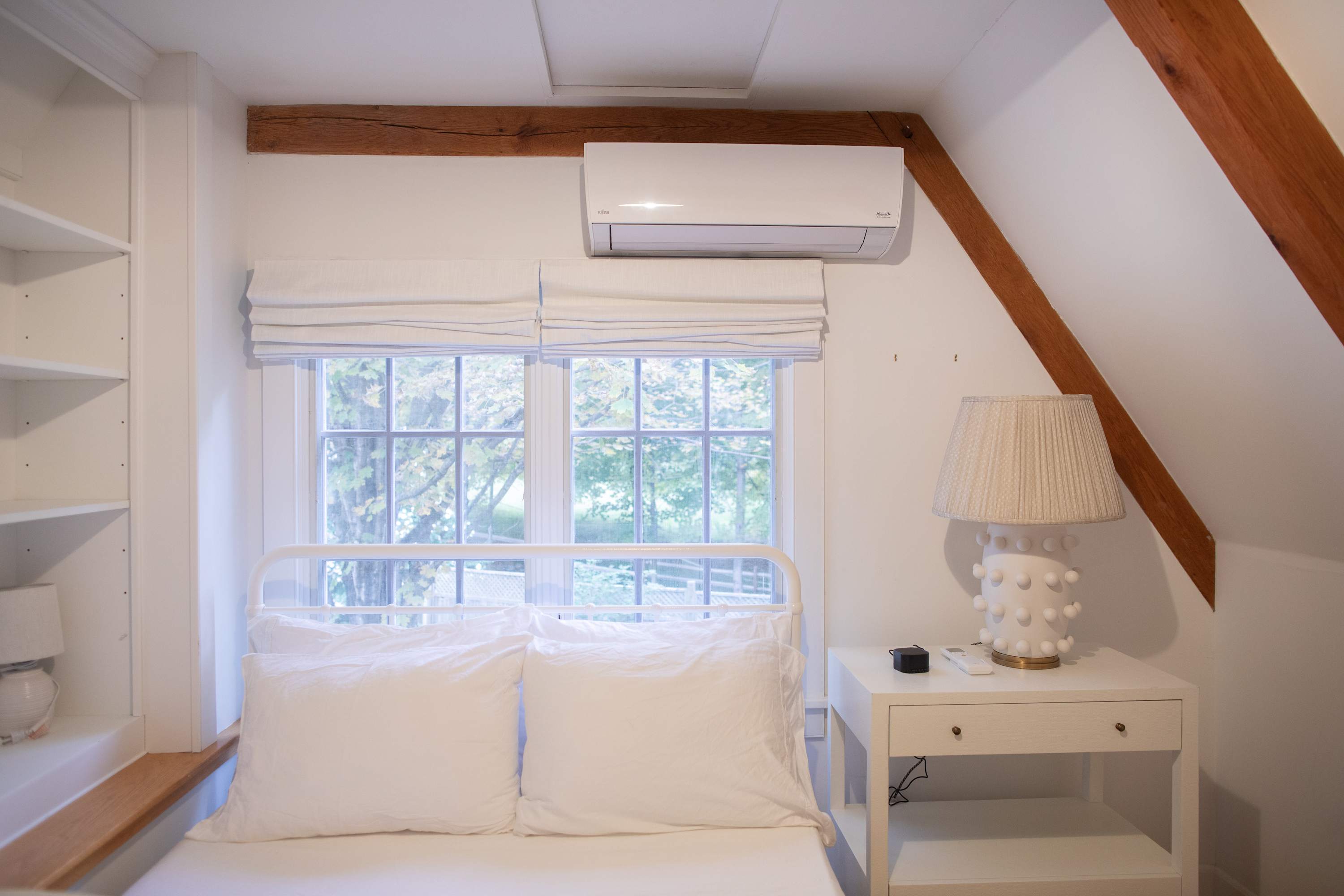 Bright bedroom with a white metal frame bed, white bedding, a white nightstand with a textured lamp, and a window overlooking greenery, all under an exposed wooden beam and with a wall-mounted AC unit.