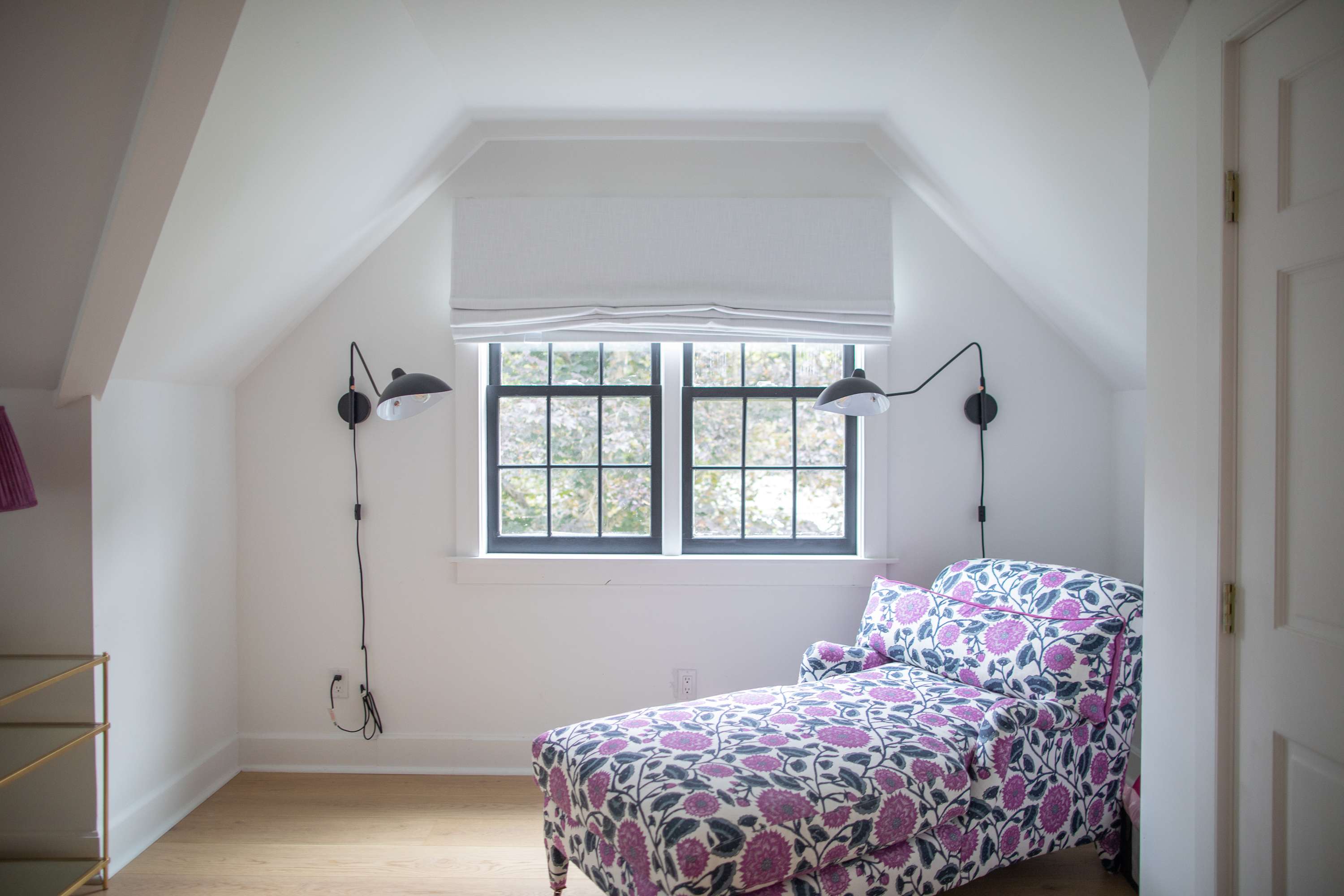 Bright room with a floral patterned chaise lounge, two black wall-mounted sconces, and a large window offering views of the outdoors, all under a sloped white ceiling.