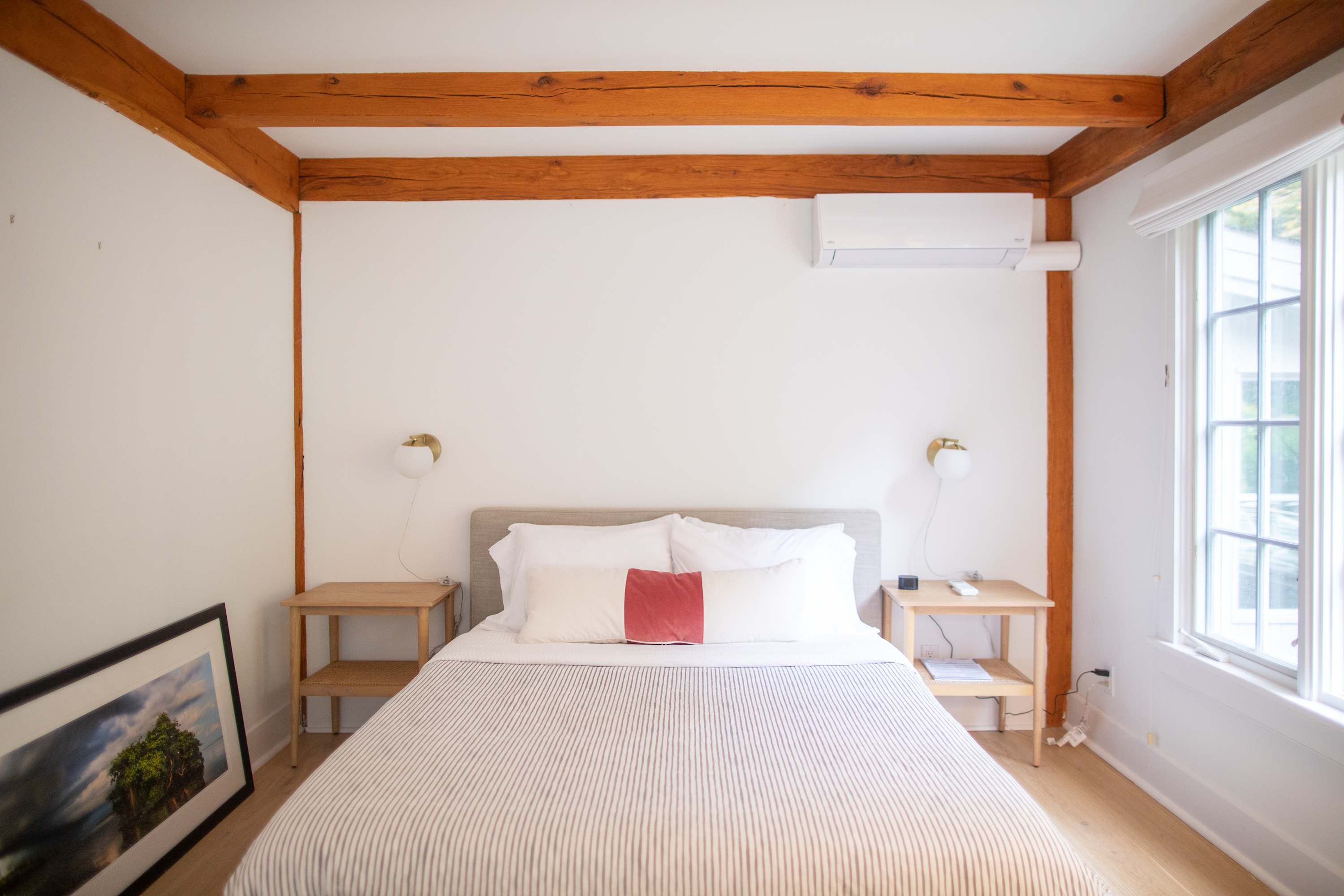 A bright bedroom with light hardwood floors and exposed wooden ceiling beams, featuring a queen-sized bed with white and red pillows. Two simple wooden nightstands with modern sconces flank the bed, and a large window provides natural light.